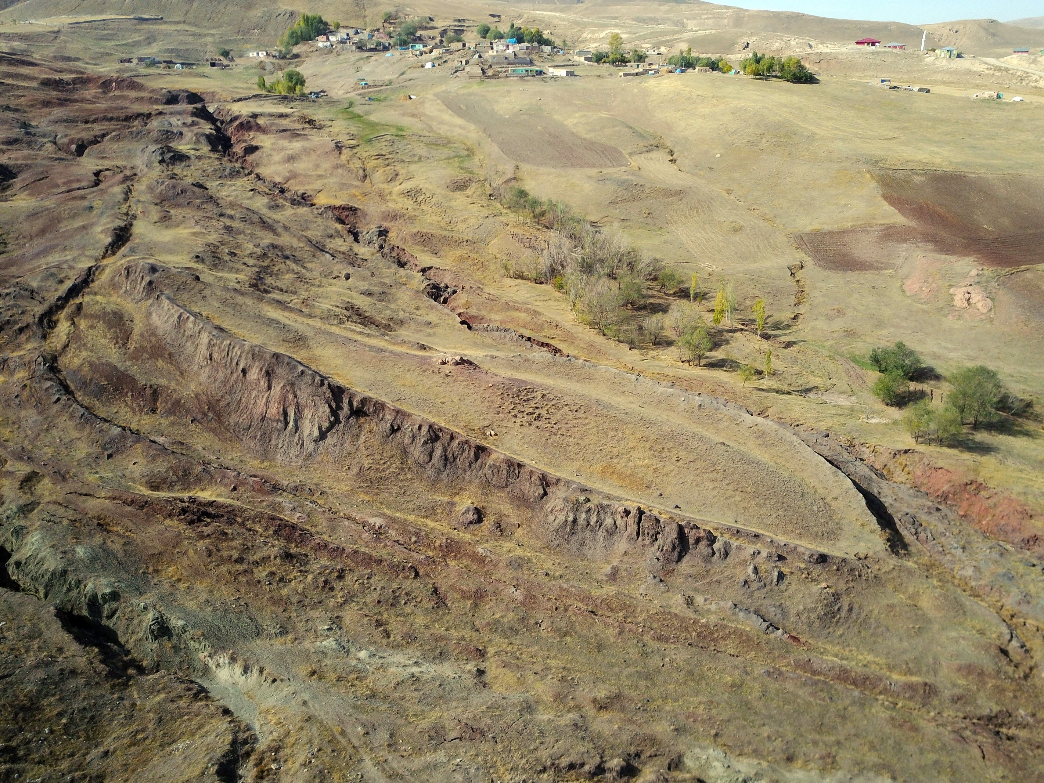 Luftaufnahme einer hügeligen, trockenen Landschaft mit Spuren auf dem Boden nahe einem Dorf.