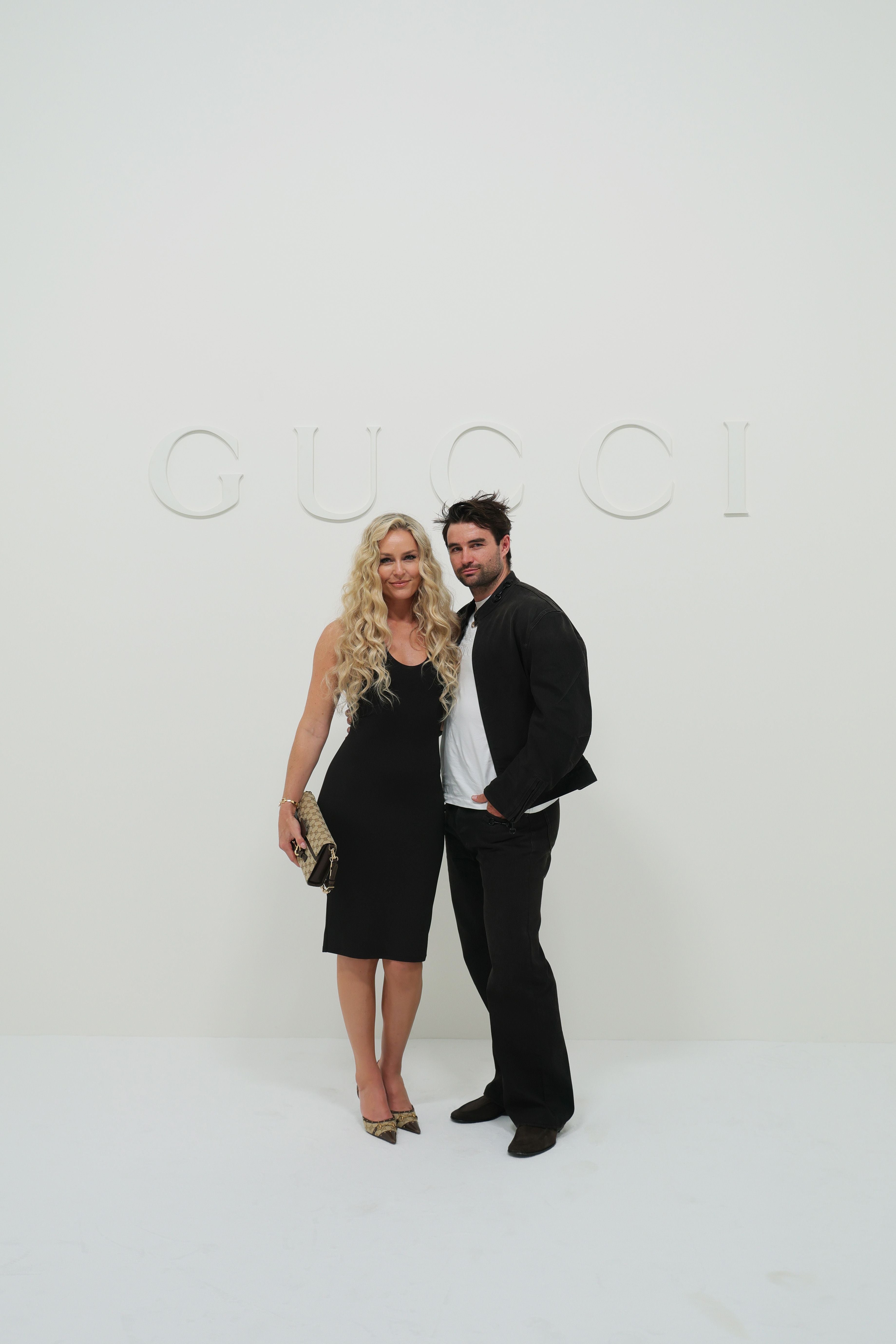 Eine Frau und ein Mann posieren schick gekleidet vor einer weißen Wand mit Gucci-Schriftzug.