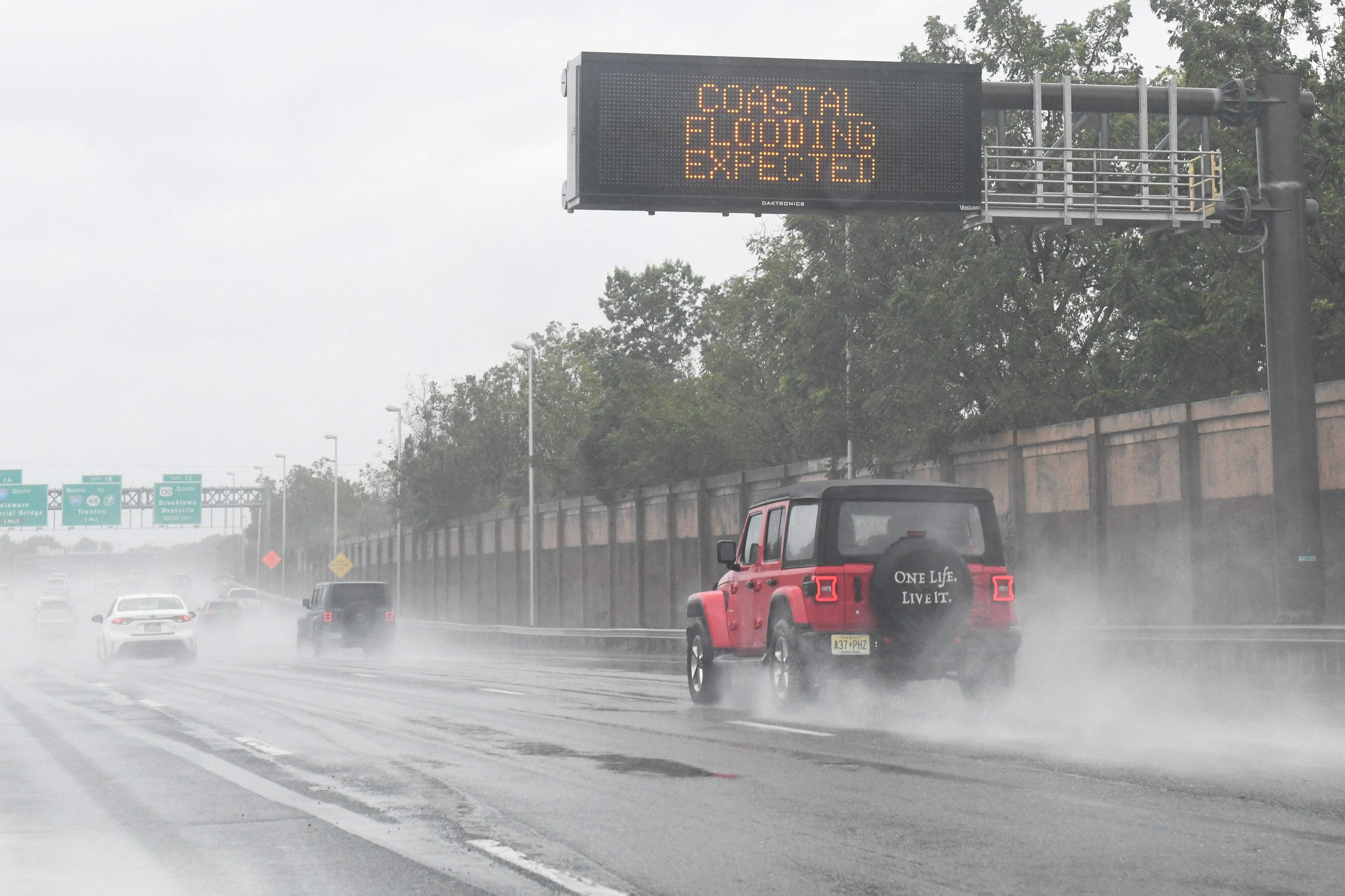 Autos fahren bei starkem Regen auf einer Autobahn, ein Schild warnt vor Küstenüberschwemmungen.