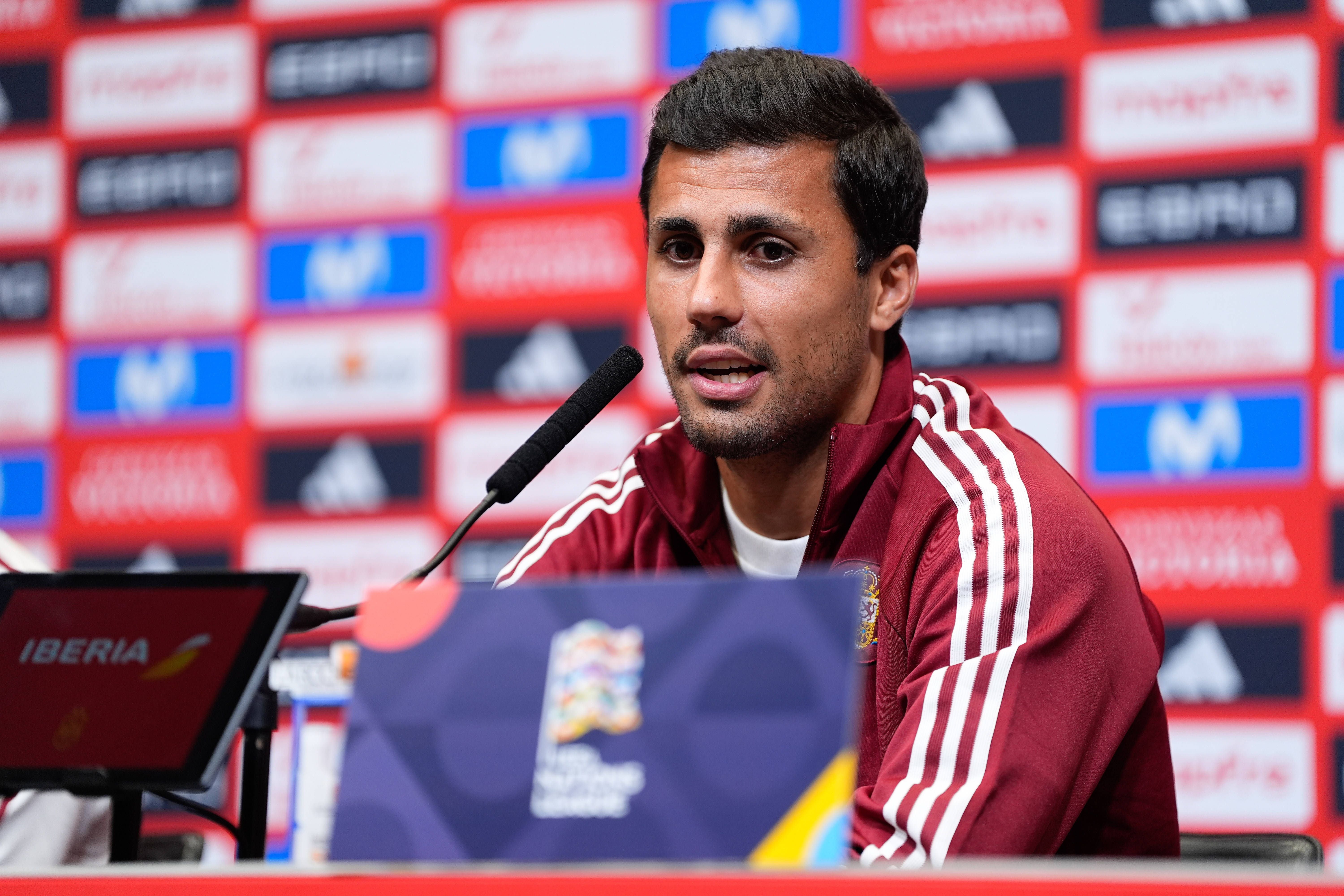 Ein spanischer Fußballspieler spricht bei einer Pressekonferenz vor Sponsorenwänden ins Mikrofon.