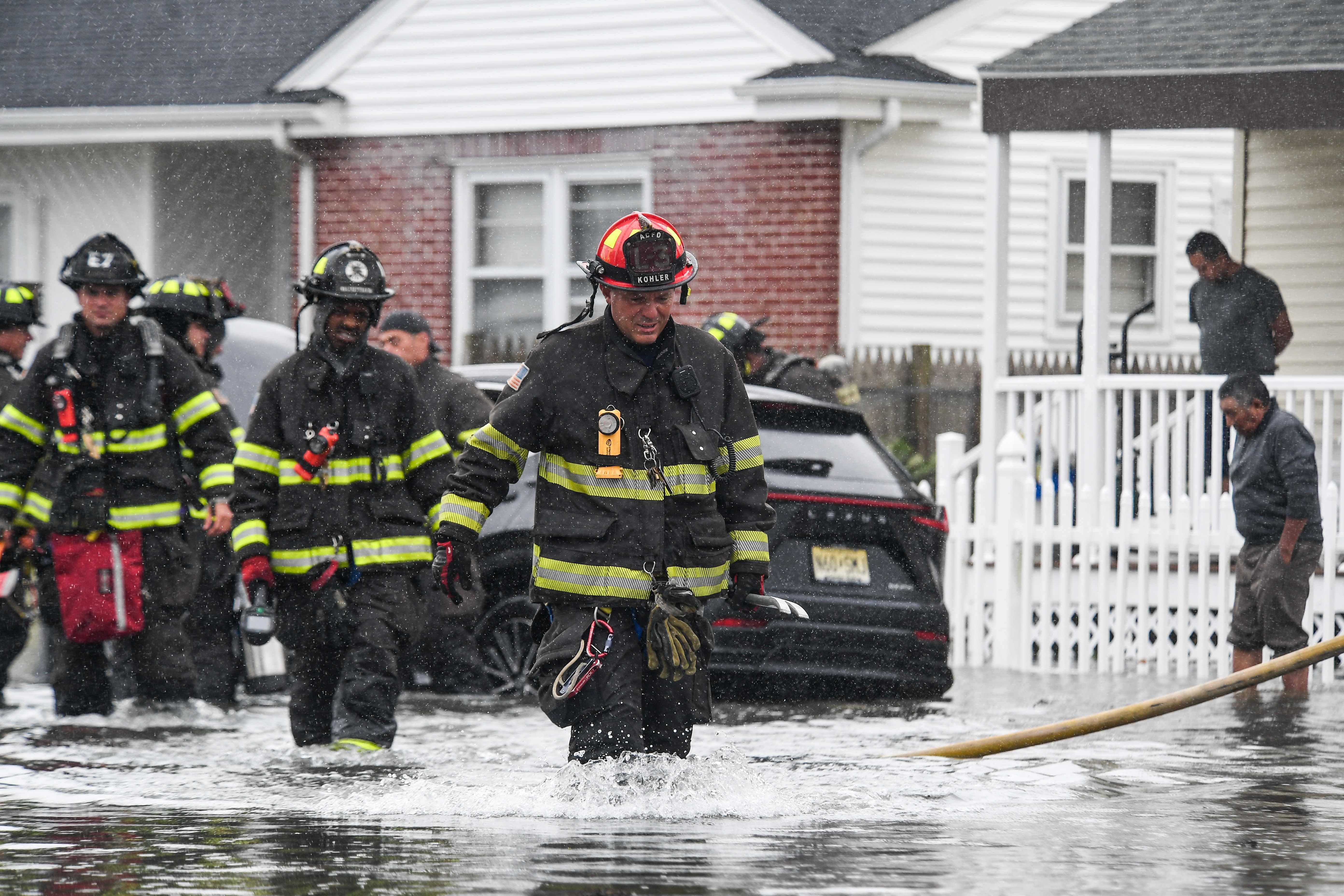 Feuerwehrleute waten bei starkem Regen durch kniehohes Hochwasser vor Wohnhäusern.
