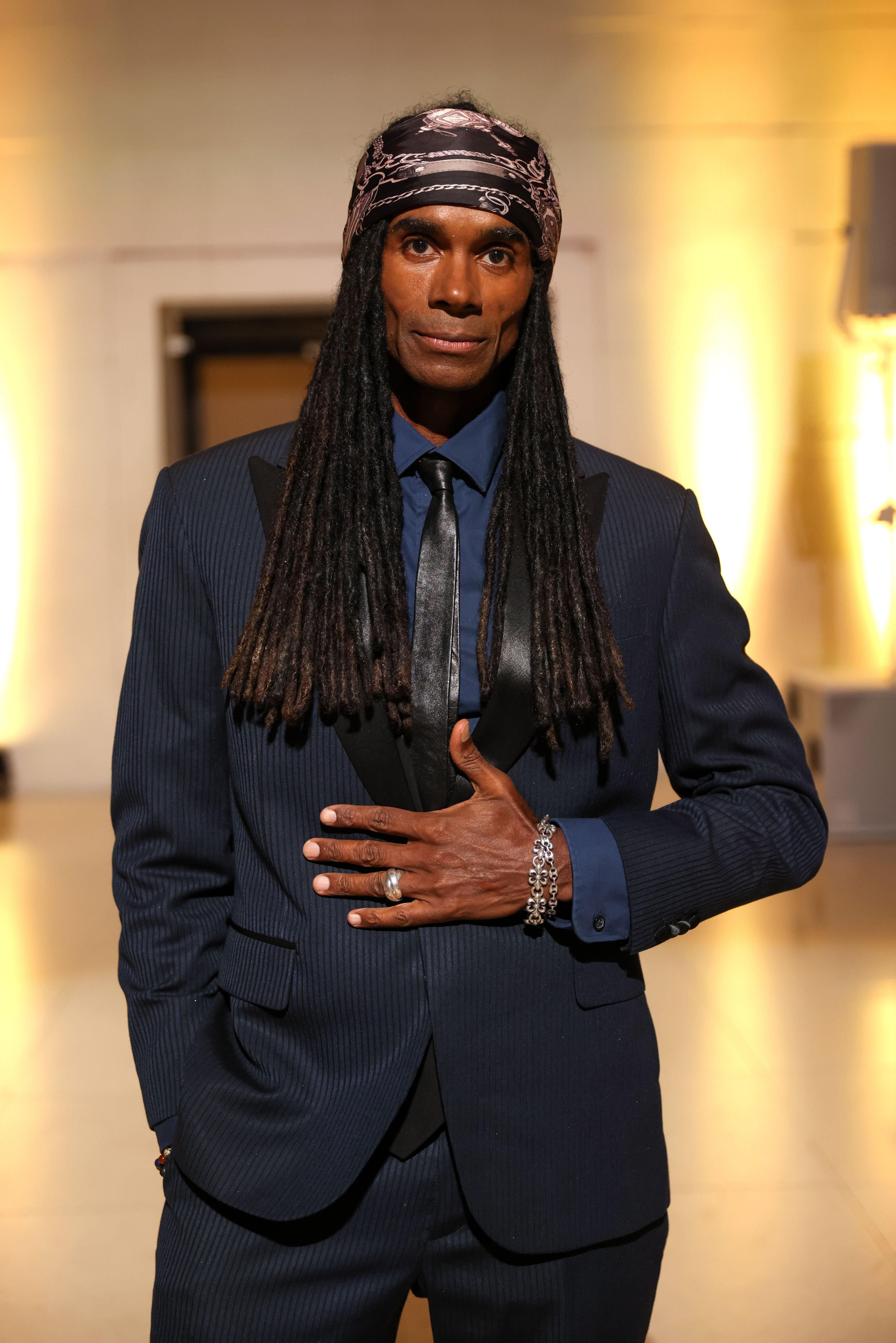 Ein Mann mit Dreadlocks, Anzug und Kopftuch steht vor beleuchtetem Hintergrund.