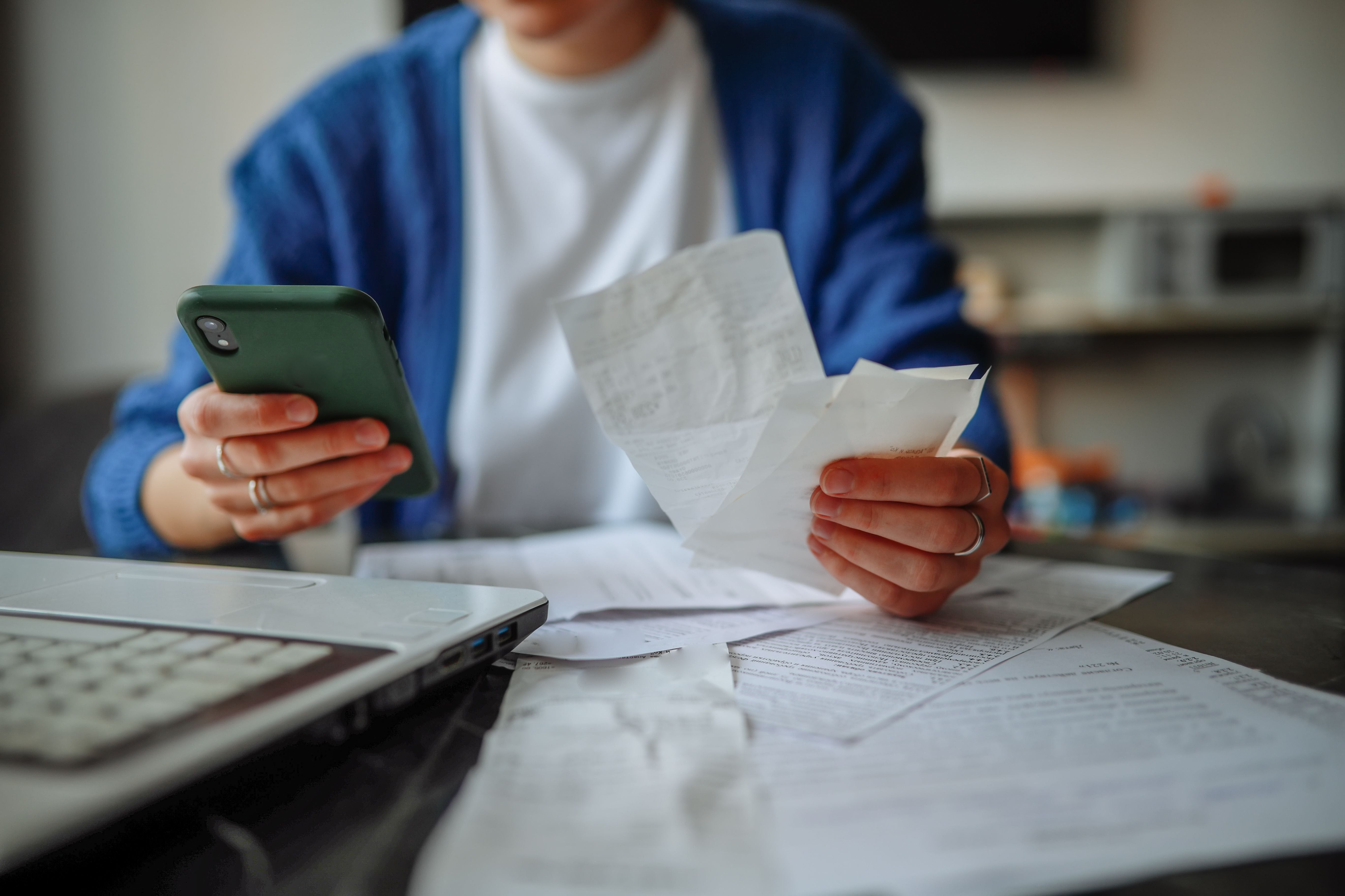 Frau sitzt mit Smartphone, Laptop und Quittungen an einem Tisch und prüft ihre Unterlagen.