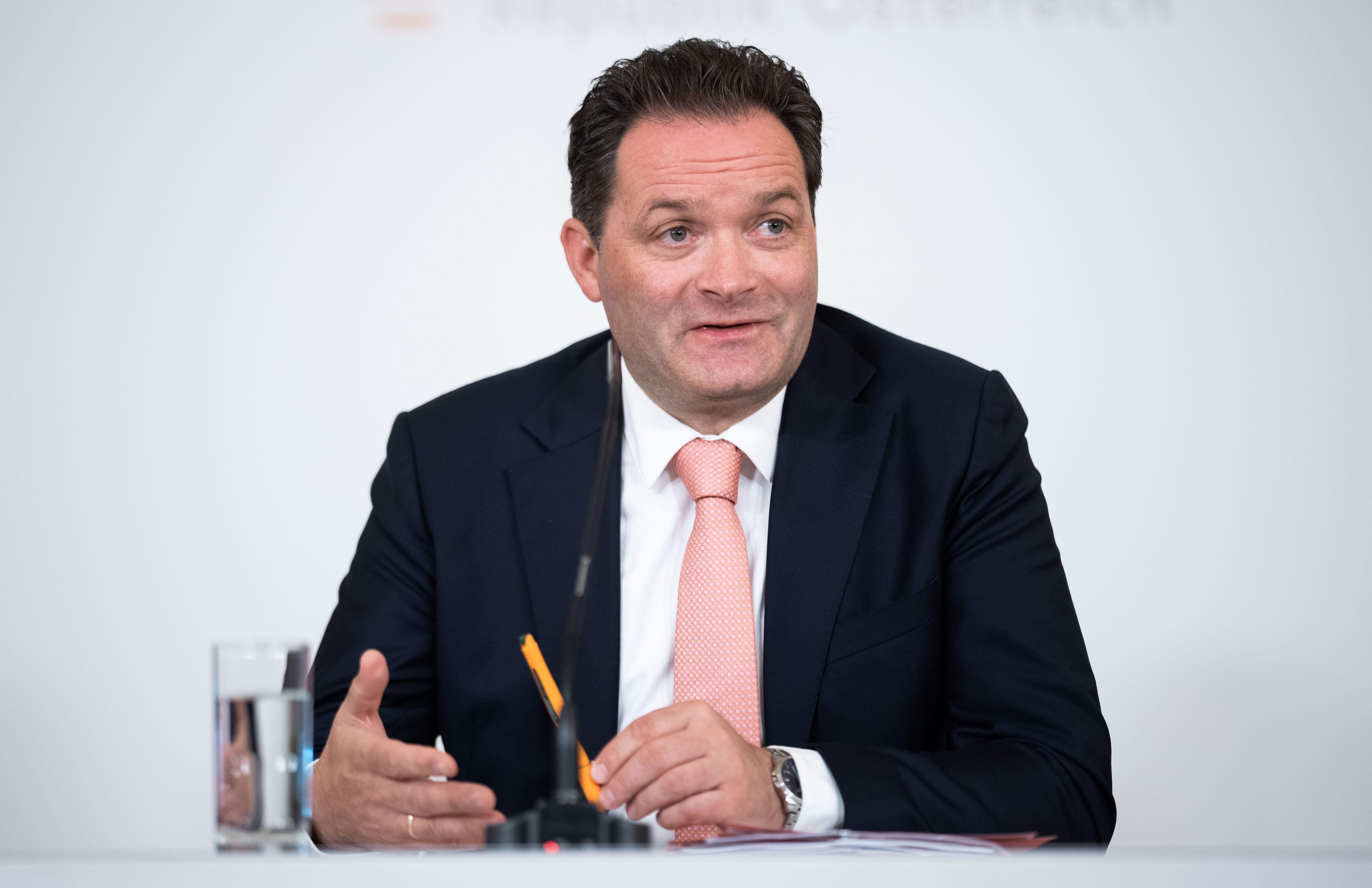 Ein Mann im Anzug spricht bei einer Pressekonferenz, vor ihm ein Mikrofon und ein Glas Wasser.