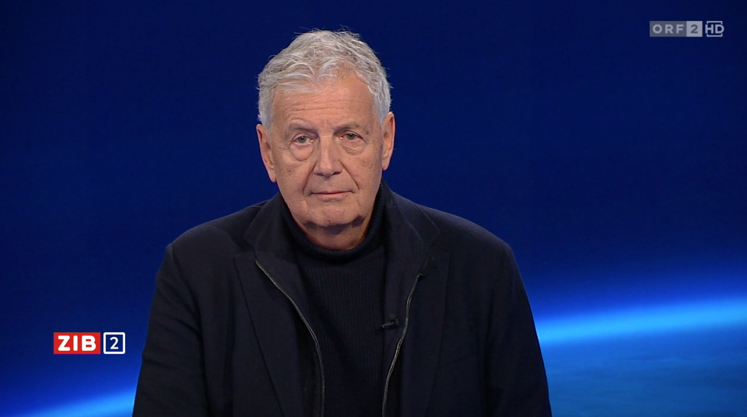 Ein älterer Mann mit grauen Haaren sitzt im Fernsehstudio vor blauem Hintergrund.