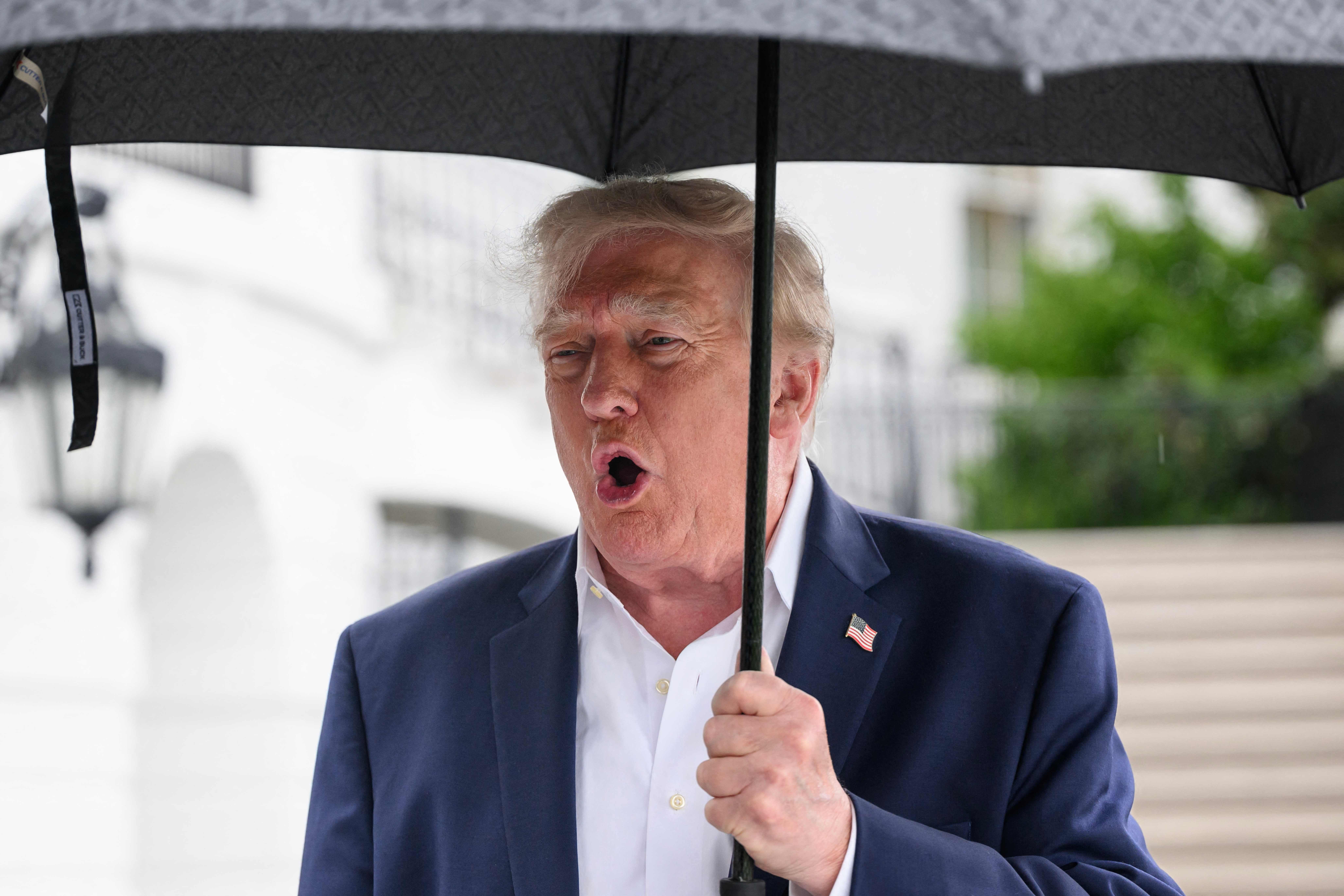 Ein Mann im Anzug mit US-Flaggenpin hält einen Regenschirm und spricht im Freien.