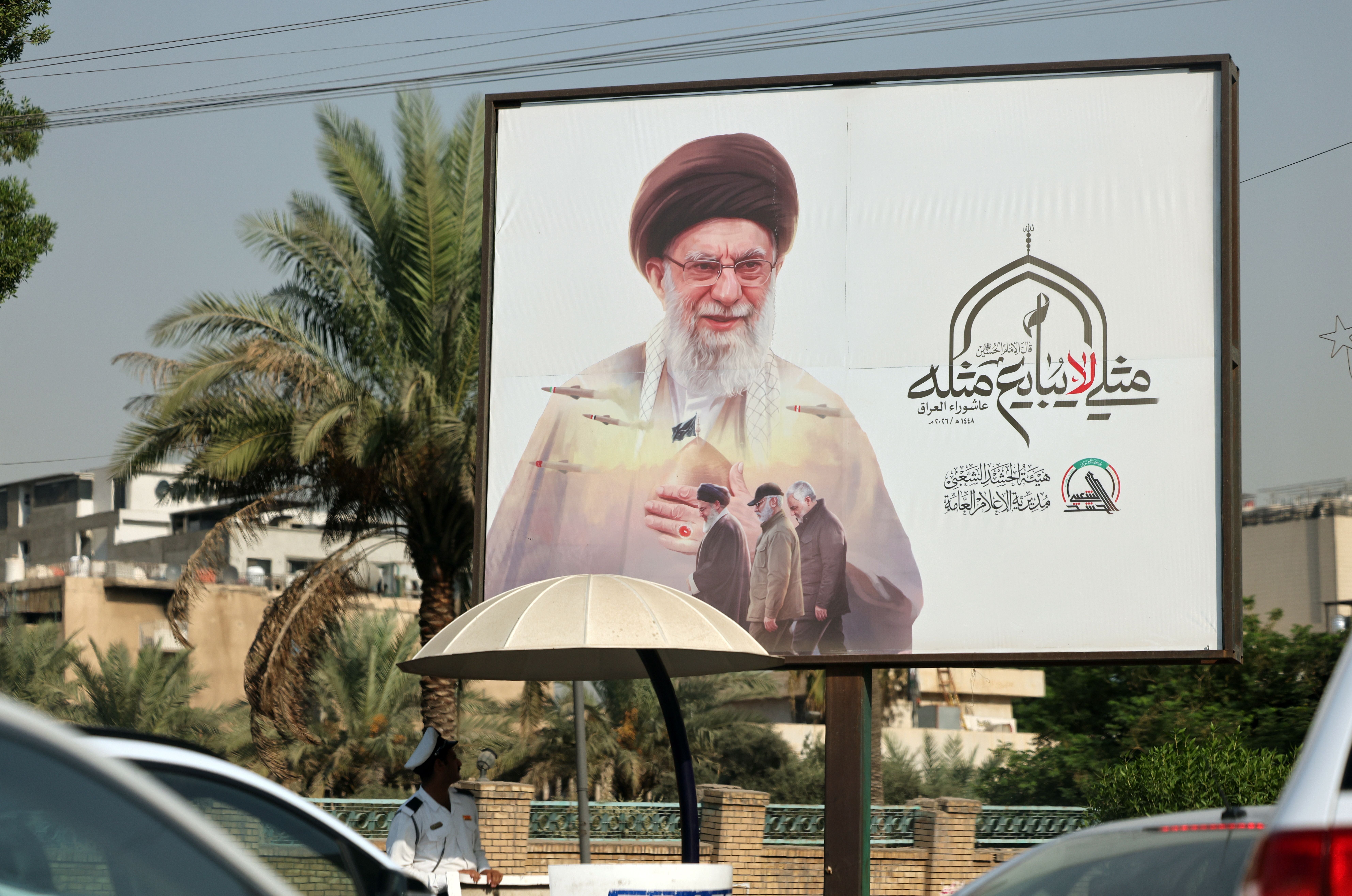 Ein irakischer Polizist steht neben einem großen Plakat mit Ali Khamenei auf einer Straße in Bagdad.