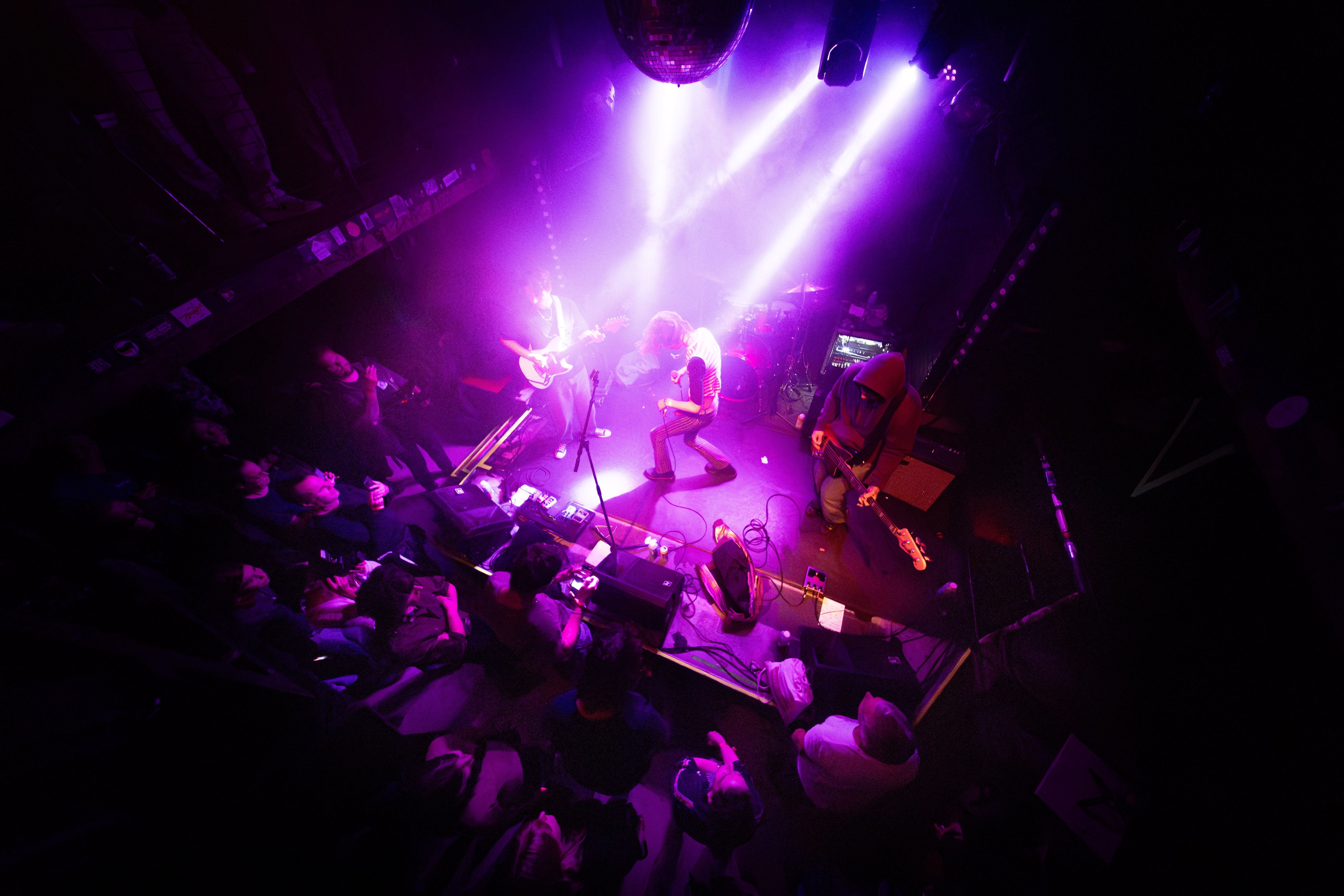 Bühnenauftritt einer Band mit Gitarren, lila Licht und jubelndem Publikum im Club.