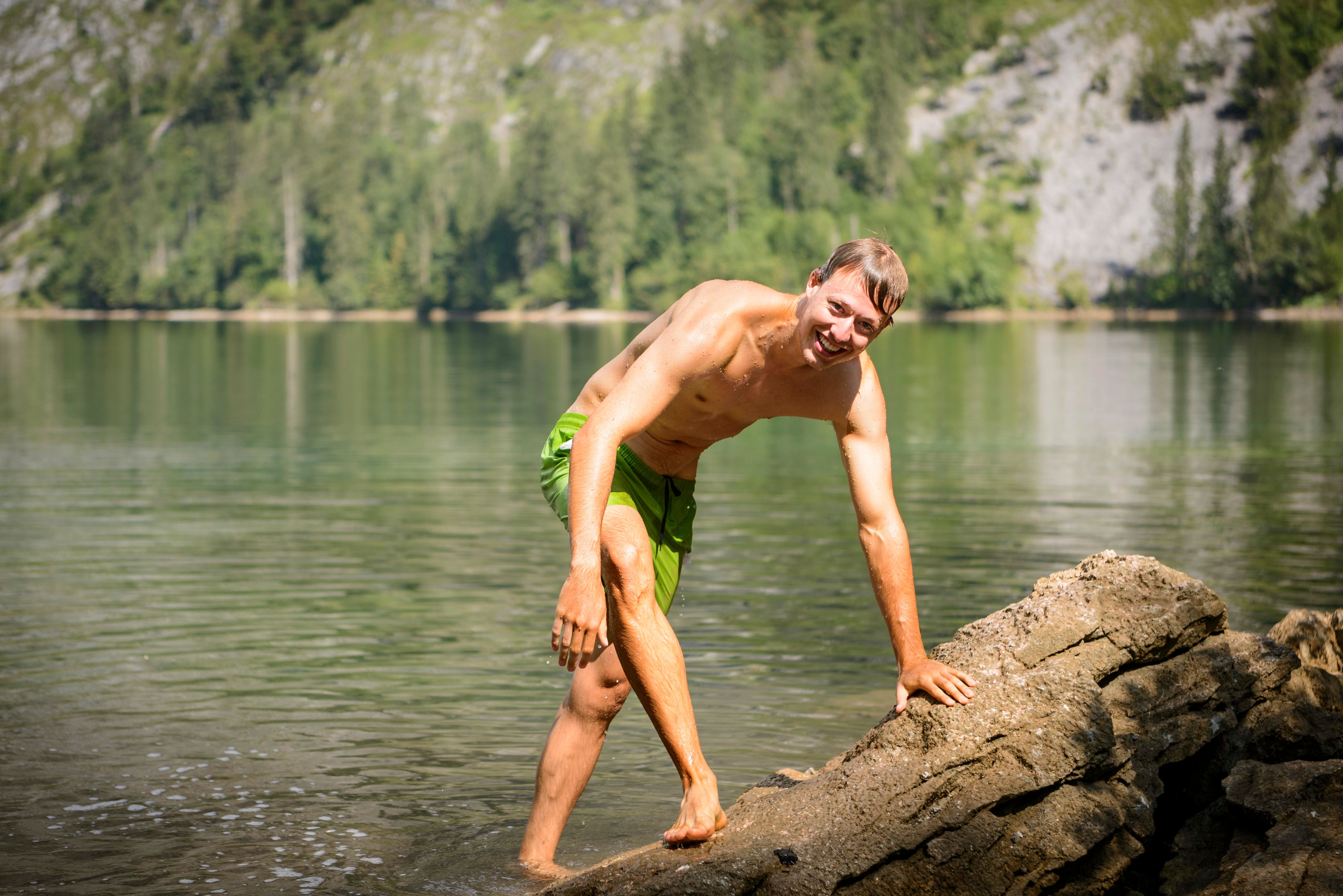 Mann in grüner Badehose steigt lachend aus einem Badesee auf einen Felsen.