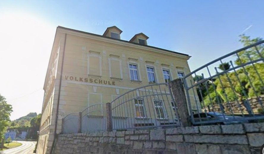 Altbau der Volksschule St. Nikola hinter einem Zaun an einer Straße bei Sonnenschein.