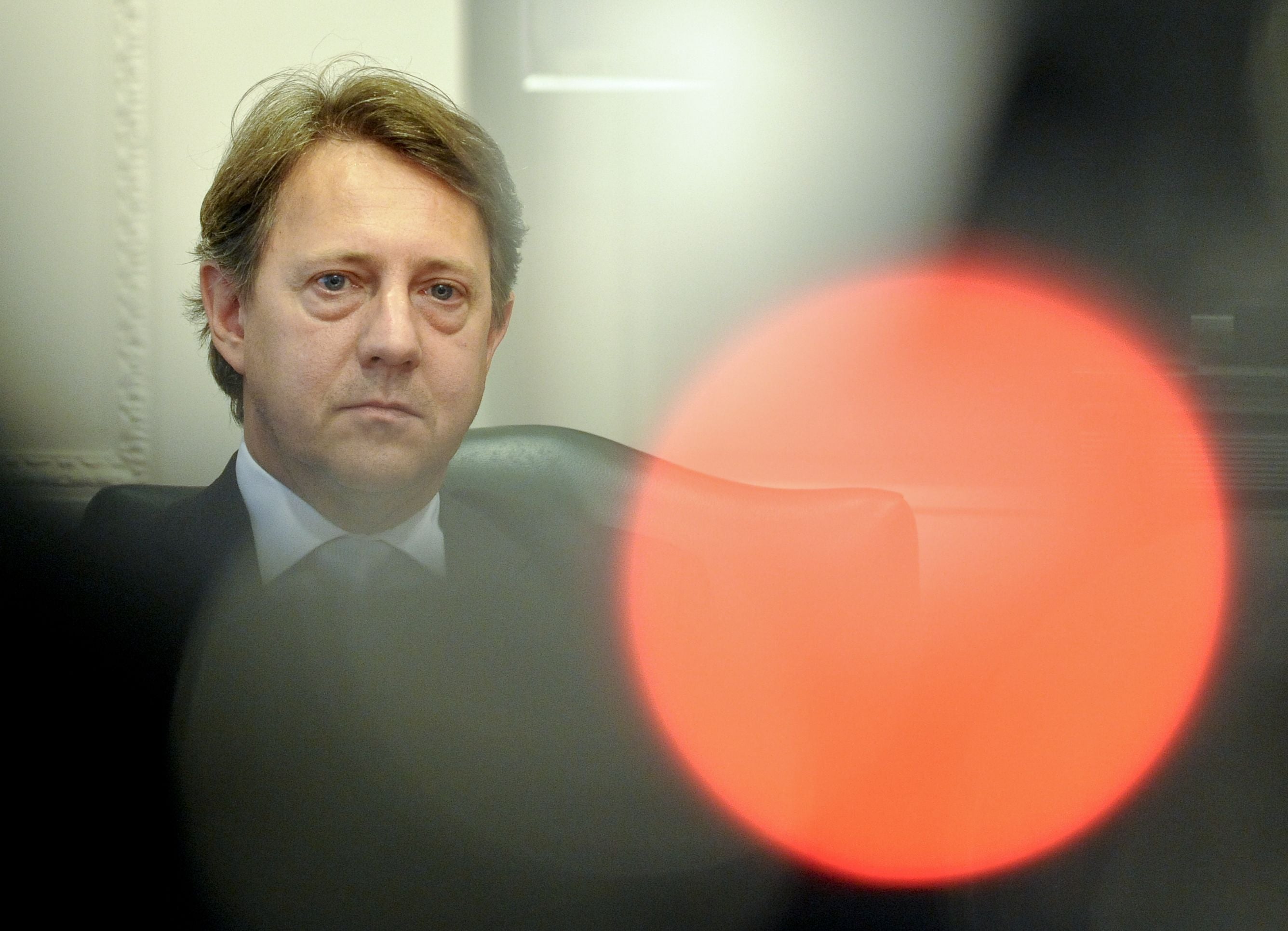 Ein Mann im Anzug sitzt ernst in einem Sessel, im Vordergrund unscharfer roter Lichtreflex.