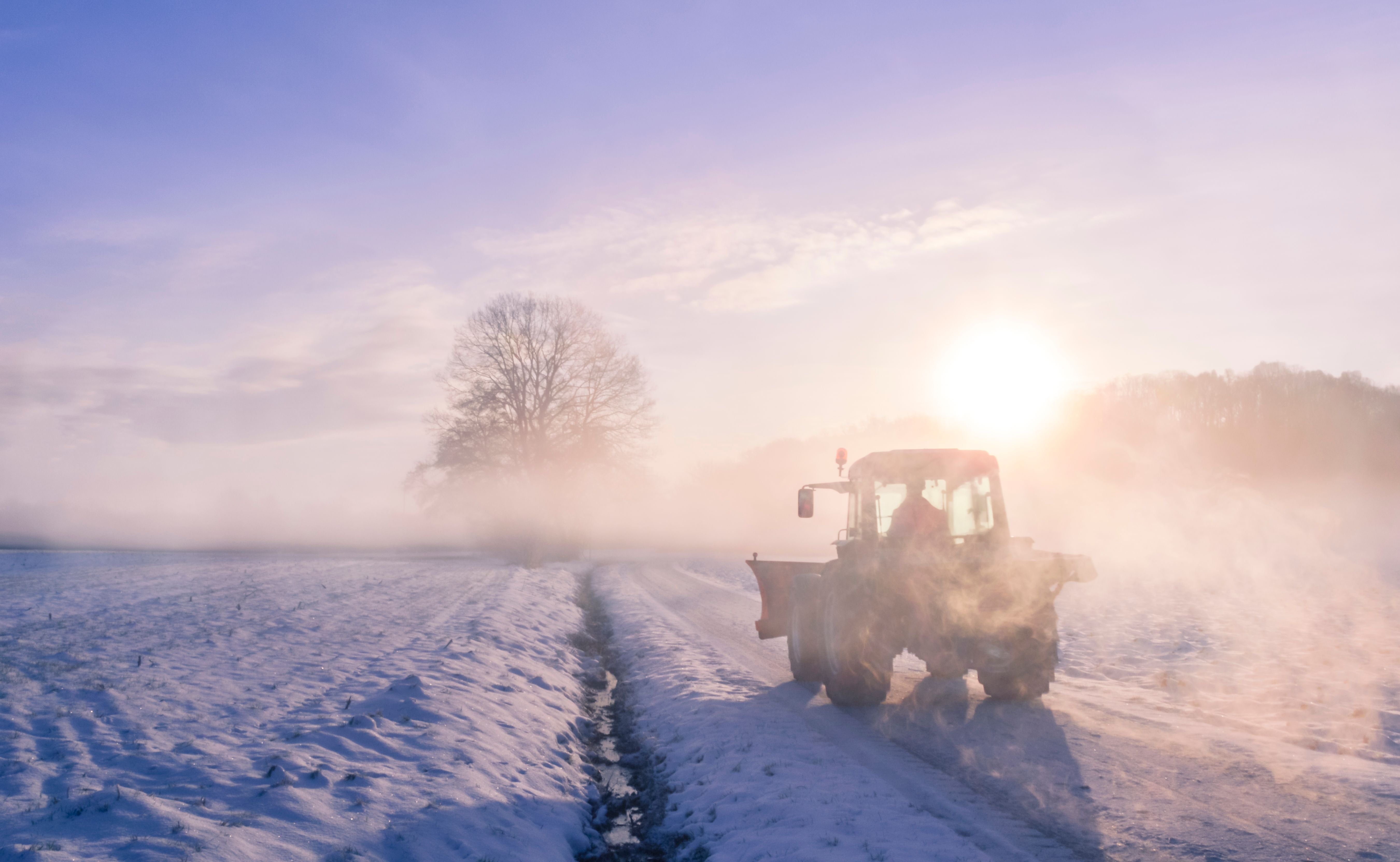 Ein Traktor fährt im Nebel über ein verschneites Feld bei Sonnenaufgang.