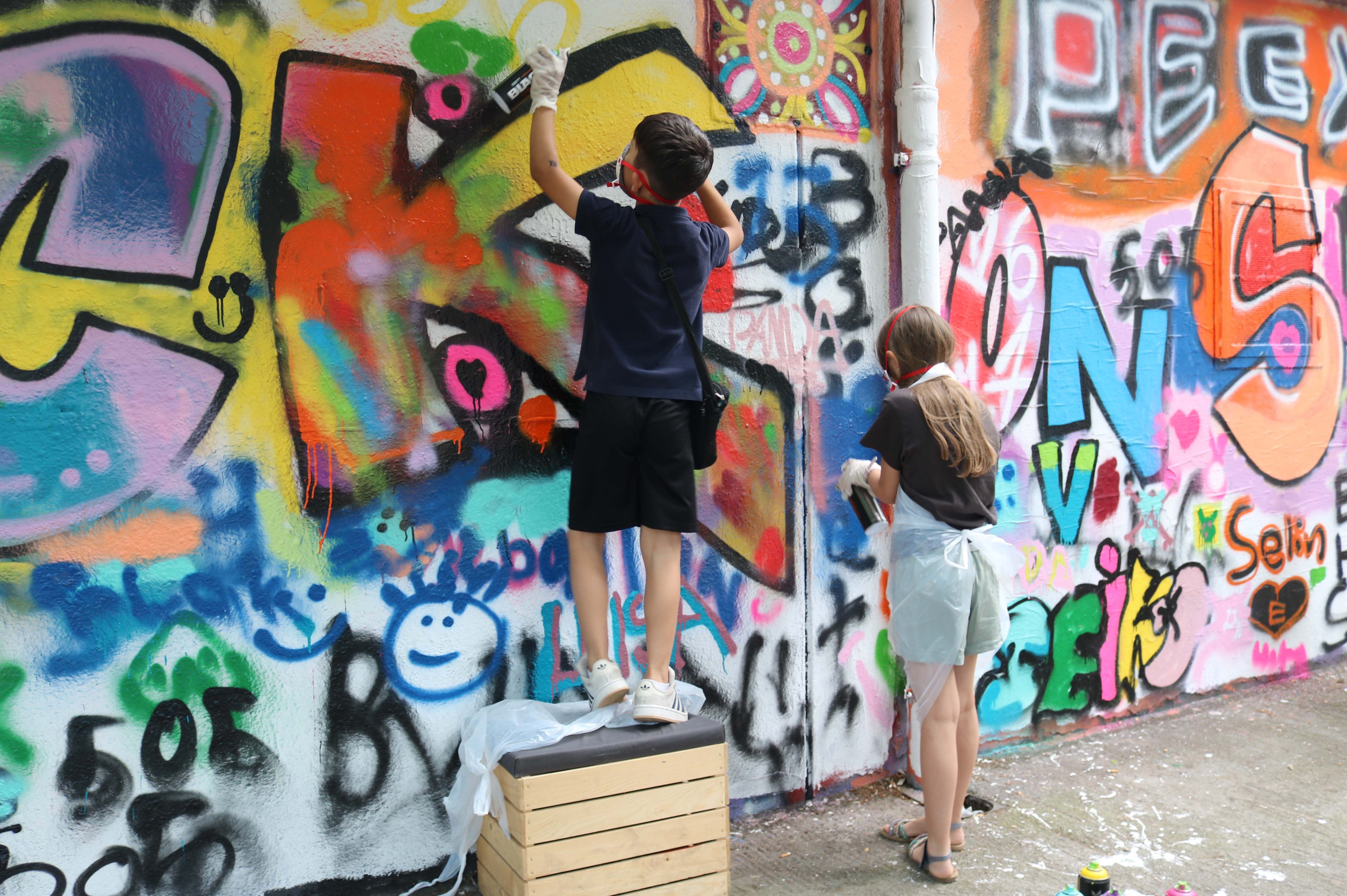 Zwei Kinder sprühen bunte Graffiti an eine Wand, eines steht auf einer Holzkiste.