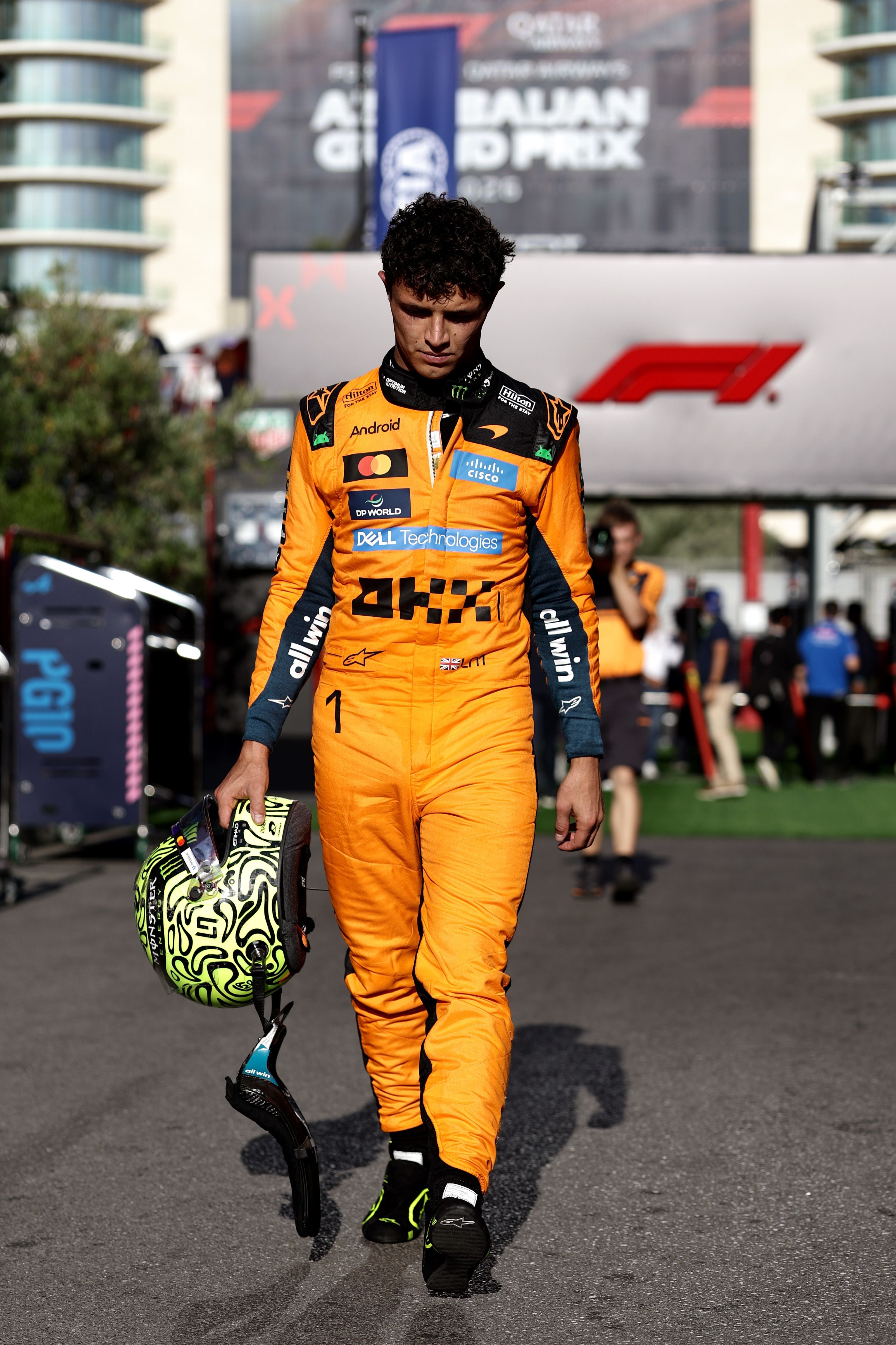 Ein Rennfahrer im orangefarbenen Overall trägt einen Helm und geht niedergeschlagen durch das Fahrerlager.