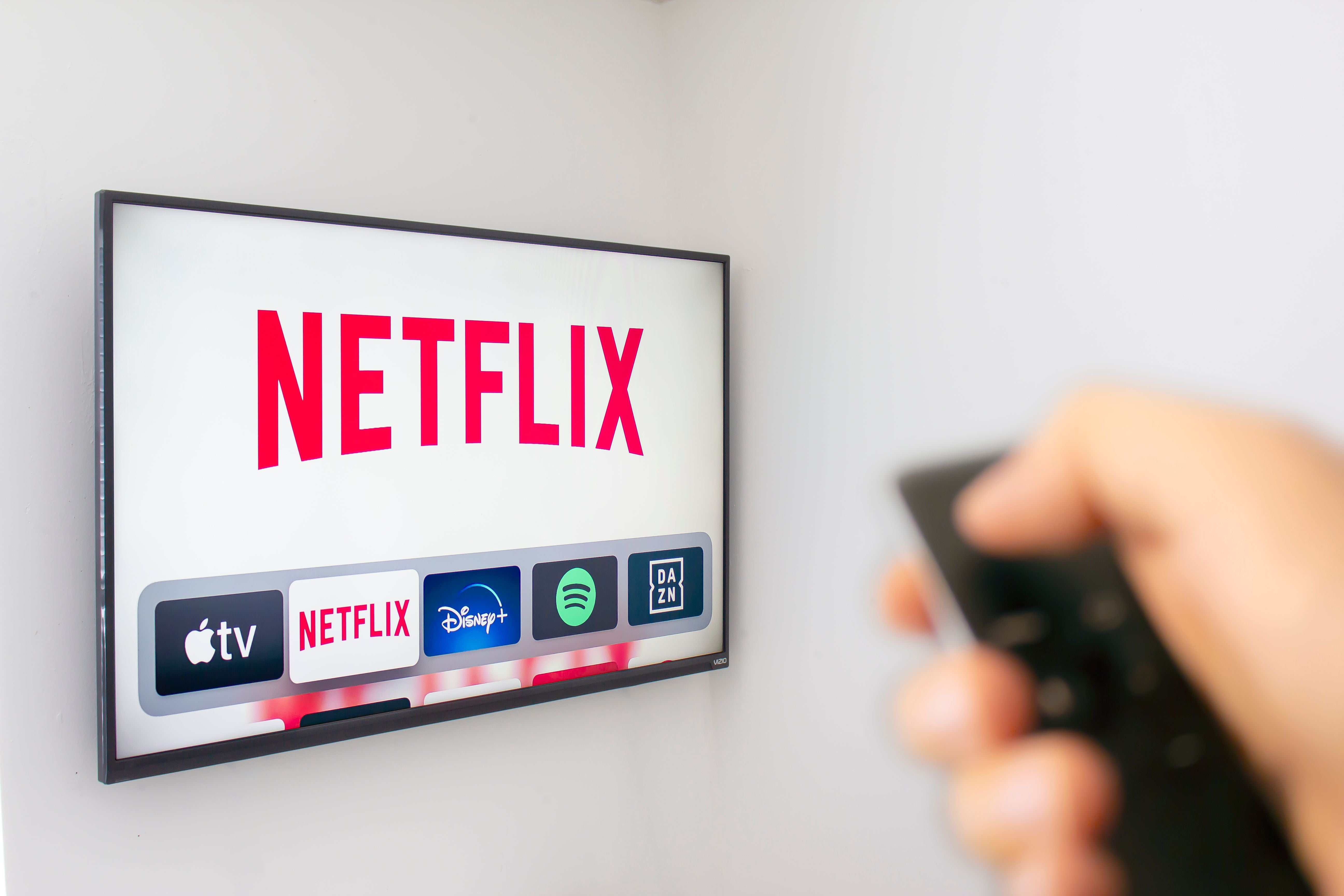 Hand hält Fernbedienung vor Fernseher mit Netflix, Apple TV, Disney+ und Spotify Apps auf dem Bildschirm.