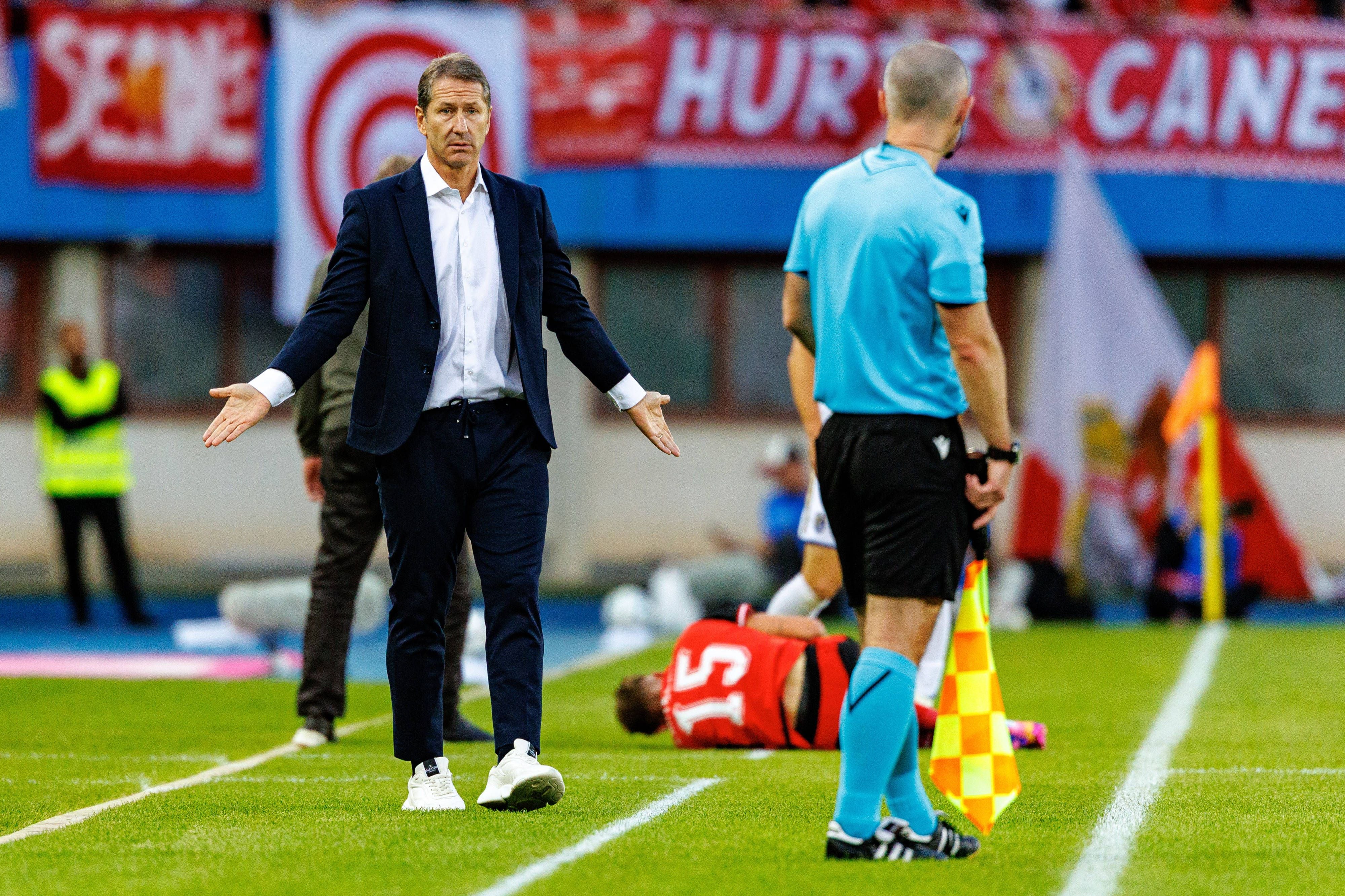 Fußballtrainer in Anzug diskutiert mit Schiedsrichter, verletzter Spieler liegt am Boden.