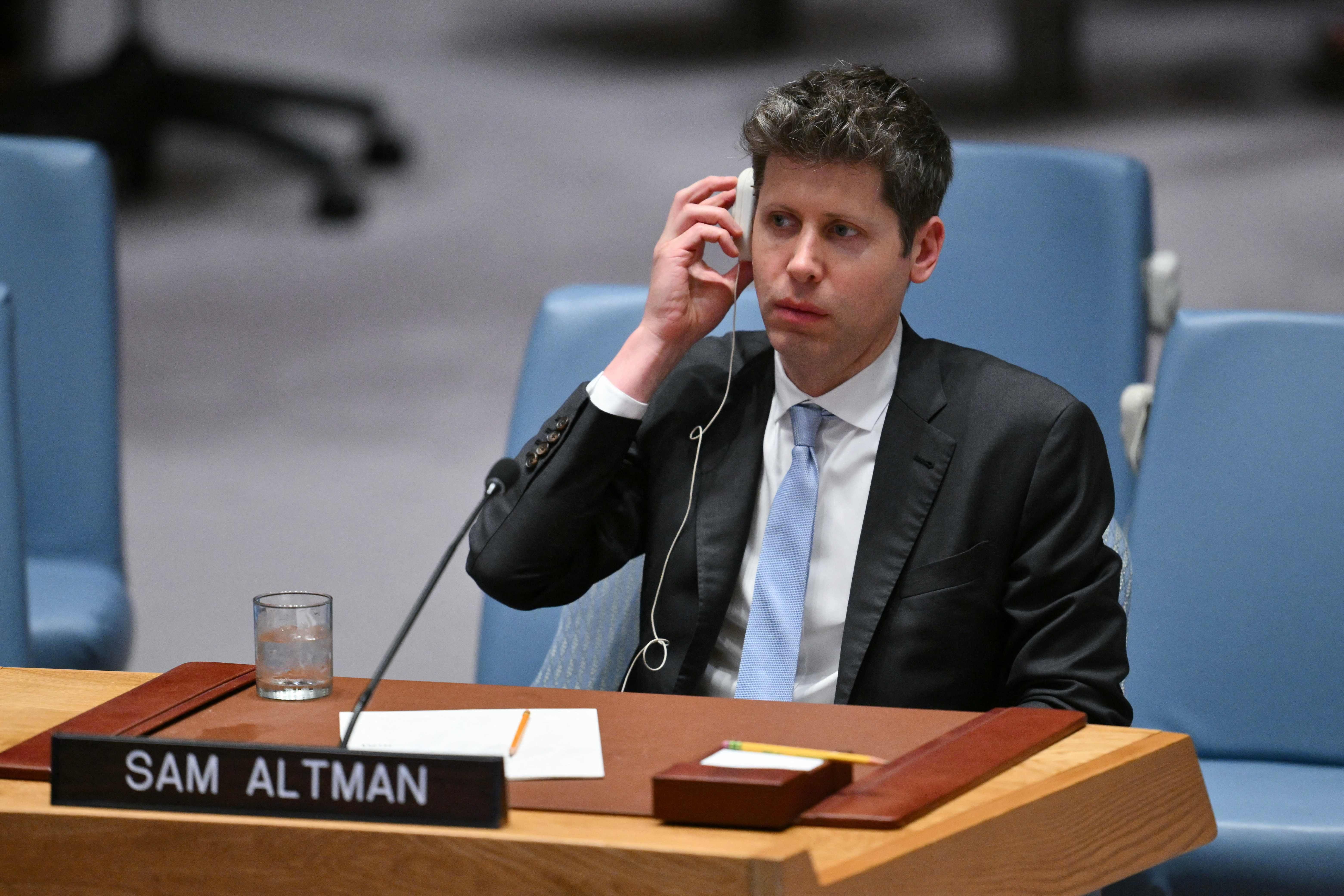 Ein Mann im Anzug sitzt an einem Tisch mit Namensschild "Sam Altman" und hält einen Kopfhörer ans Ohr.