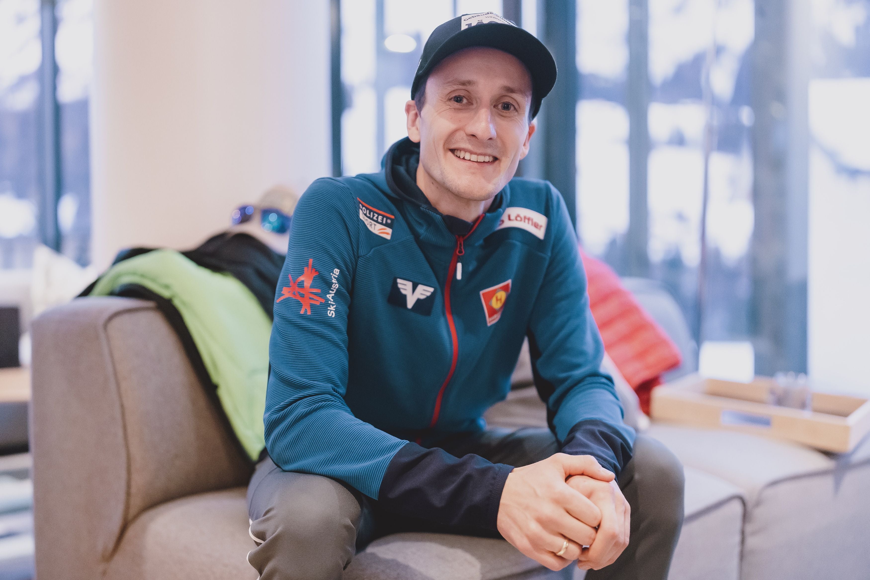 Ein Mann in Sportkleidung sitzt lächelnd auf einem Sofa in einem hellen Raum.