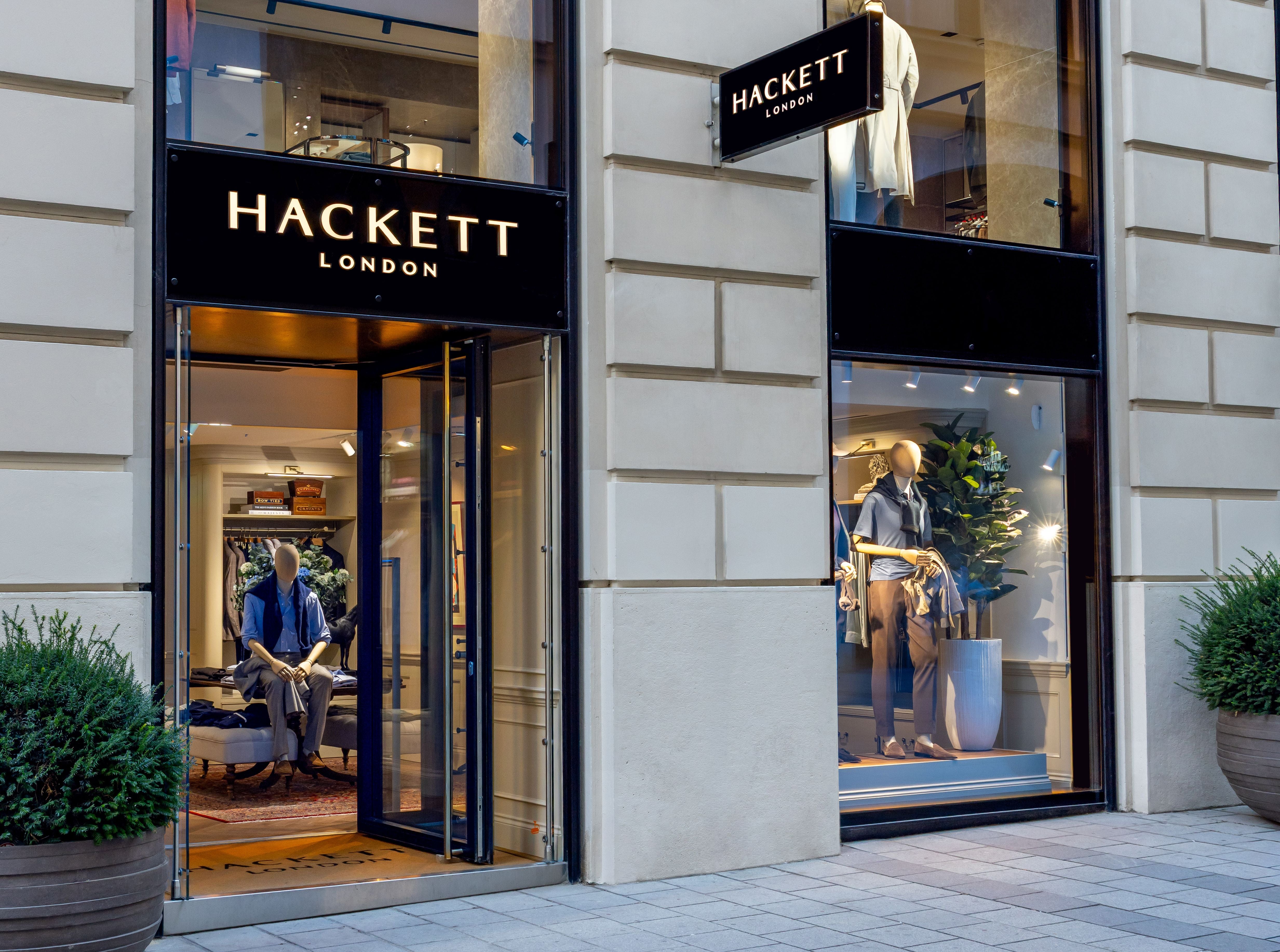 Außenansicht eines Hackett London Modegeschäfts mit Schaufensterpuppen und Pflanzen.