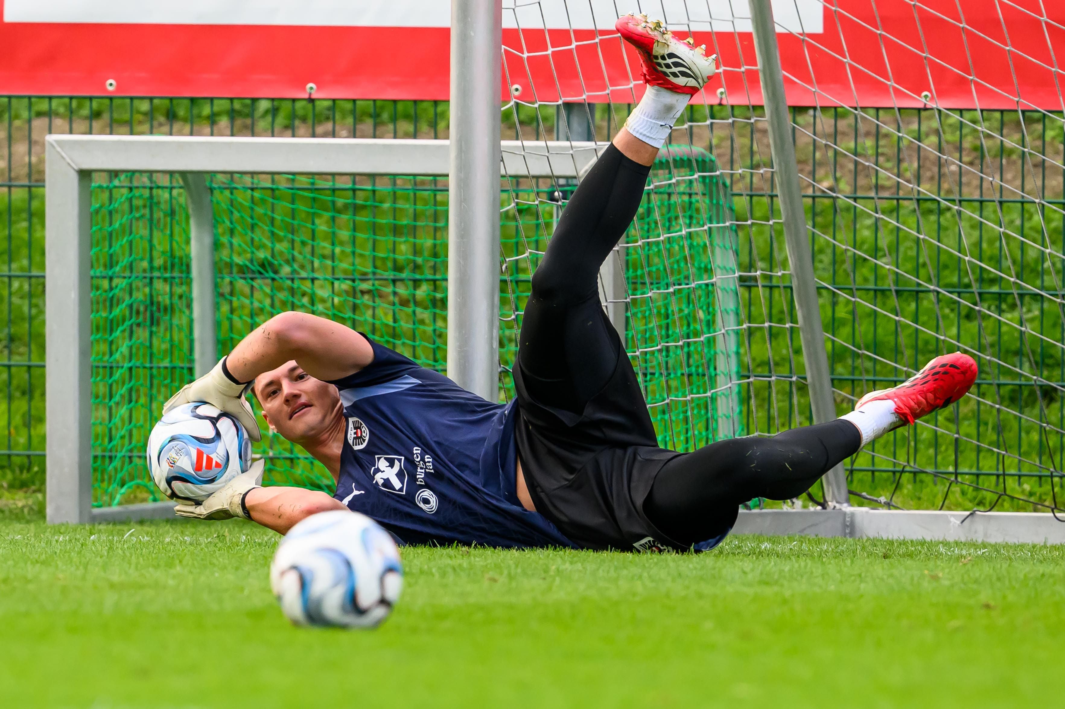 Ein Torwart in blauer Sportkleidung hält einen Fußball während eines Trainings am Boden fest.