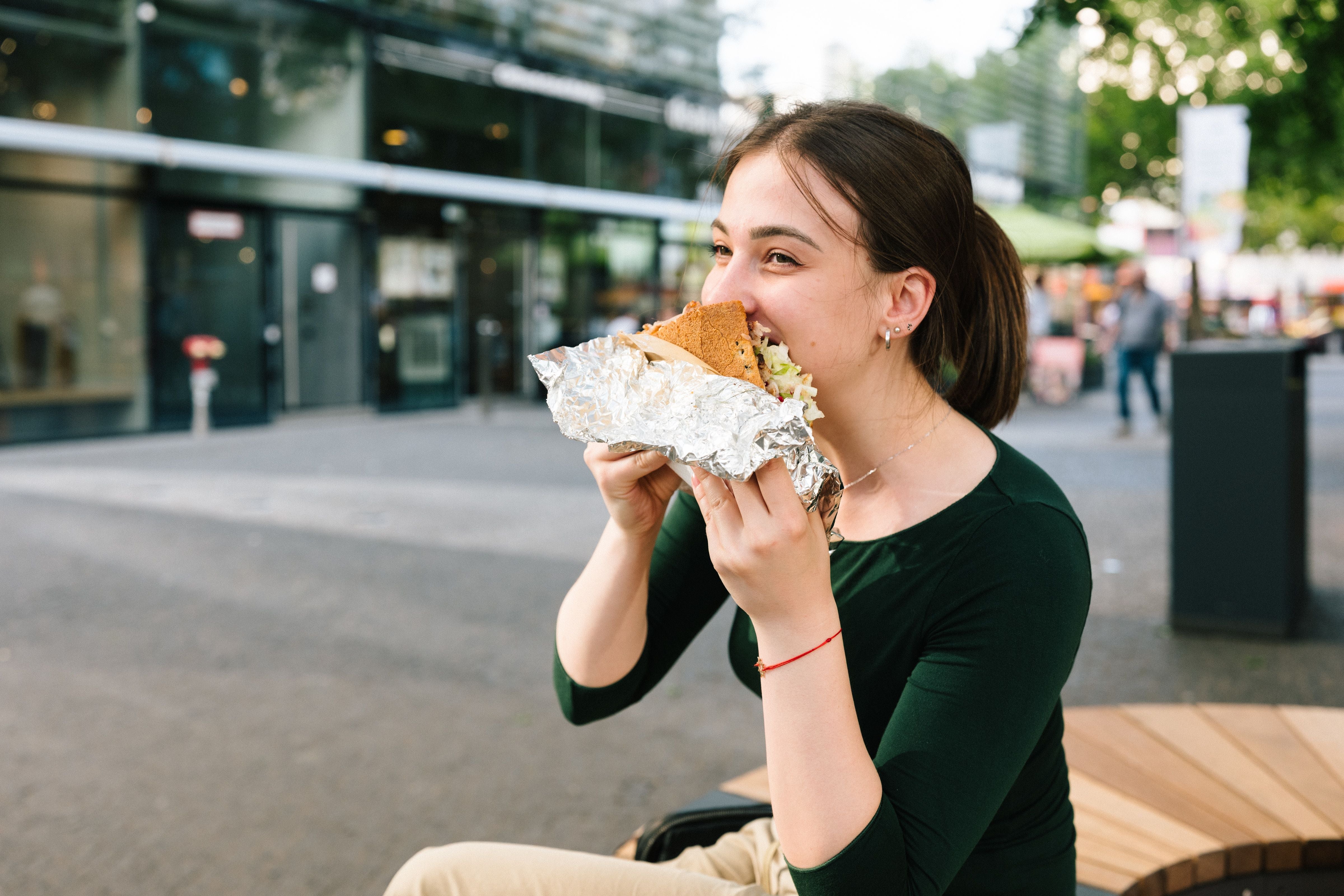Junge Frau sitzt draußen und isst einen Döner Kebab, eingewickelt in Alufolie.