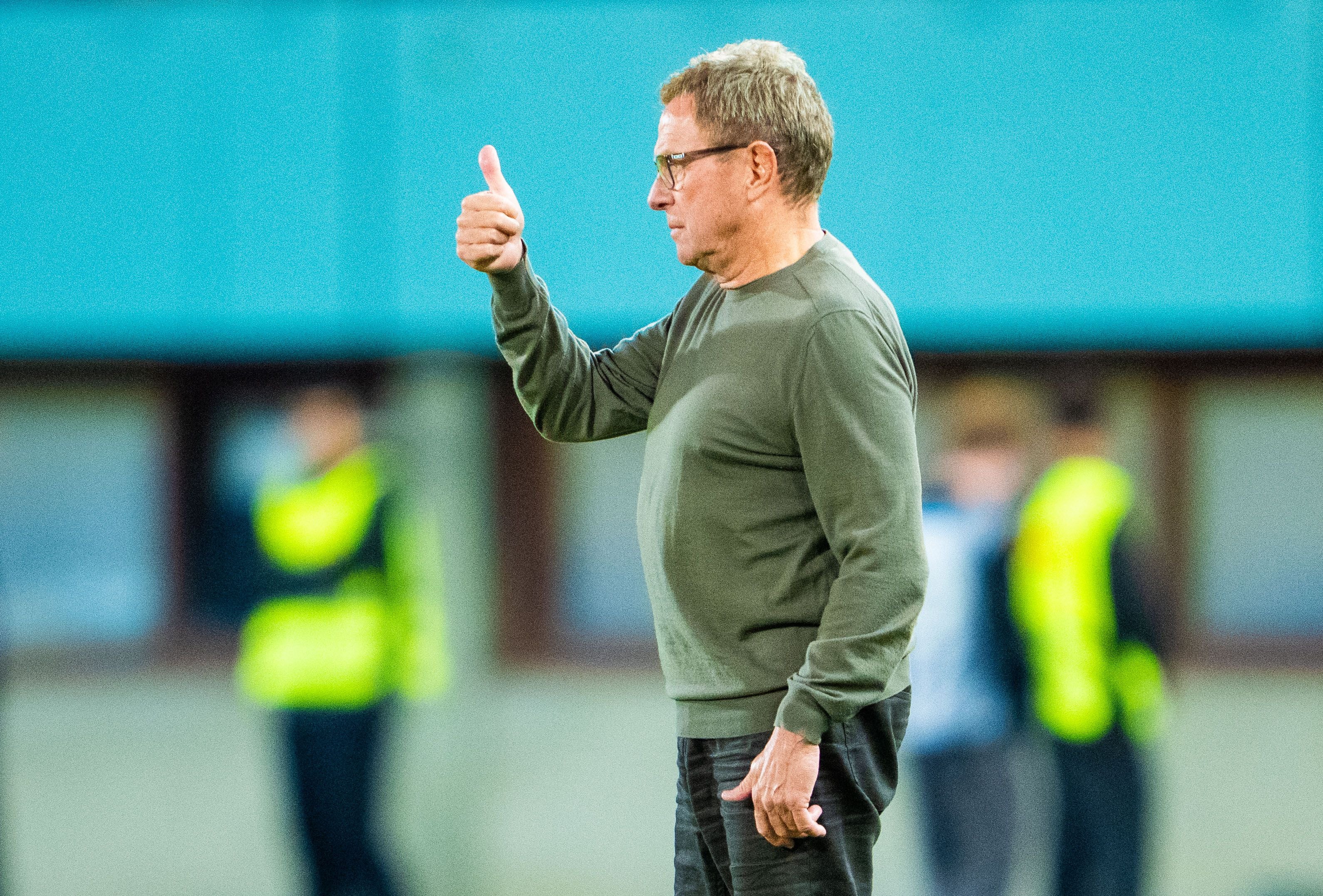 Ein Mann im grünen Pullover zeigt Daumen hoch auf einem Sportplatz.