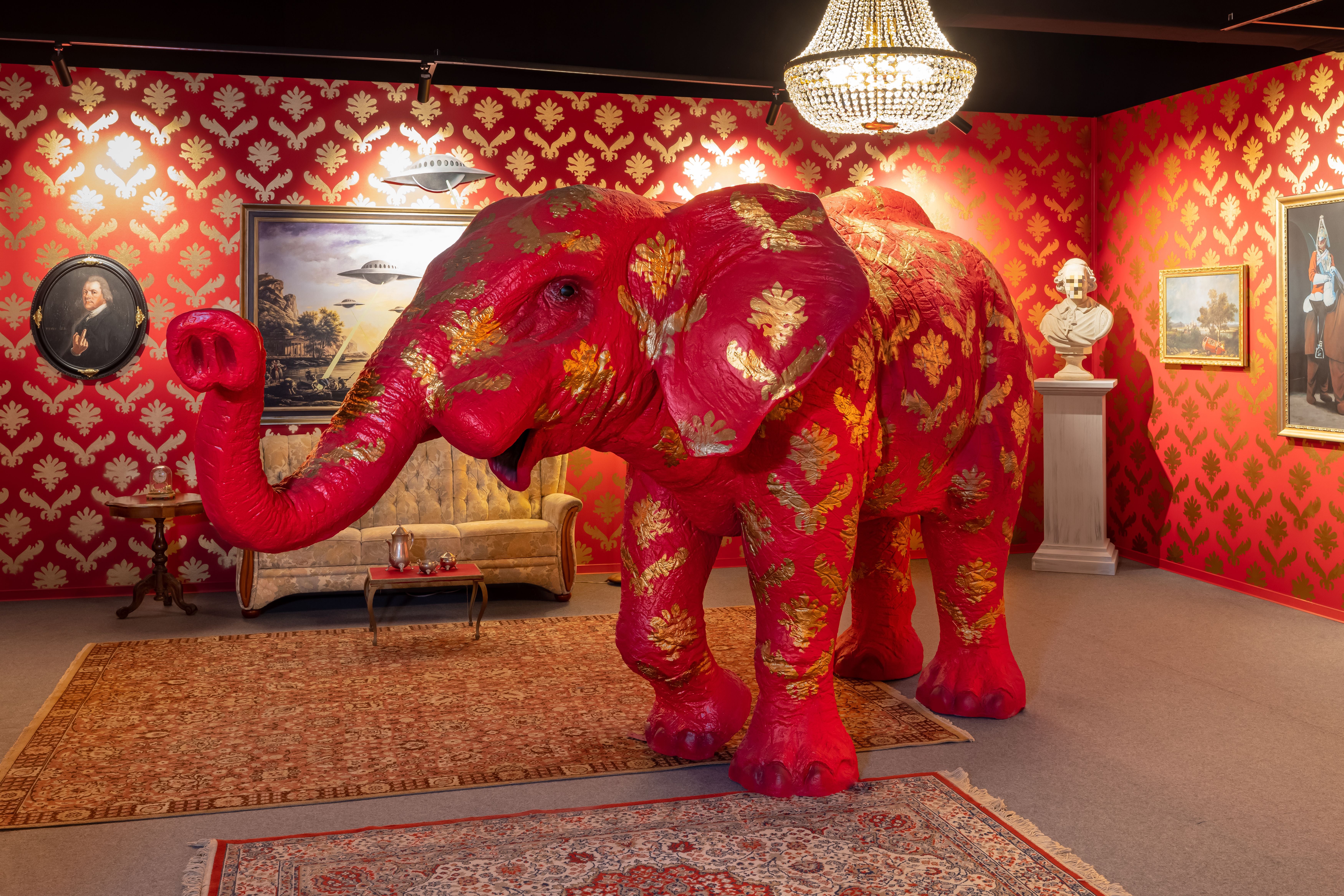 Ein großer, rot-goldener Elefant steht in einem prunkvoll dekorierten, klassisch eingerichteten Raum.