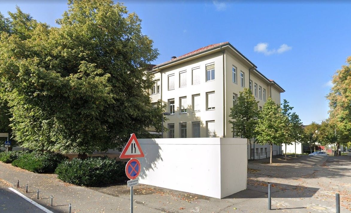 Helles Schulgebäude der Volksschule Markt Dornbirn, umgeben von Bäumen bei sonnigem Wetter.