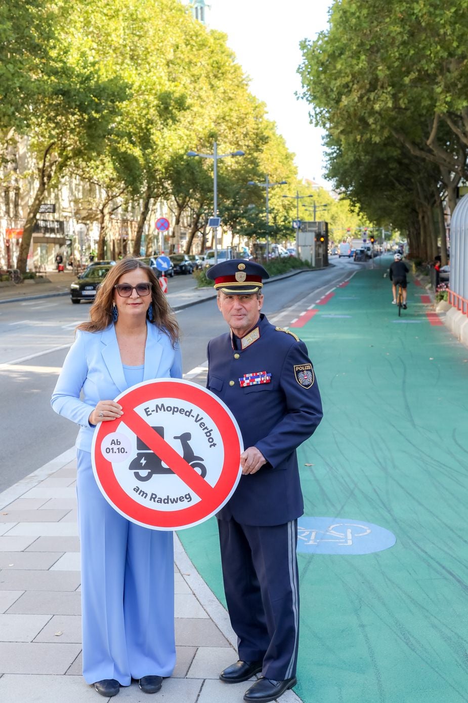 Eine Frau und ein Polizist halten ein Schild „E-Moped-Verbot am Radweg“ auf einem Radweg.
