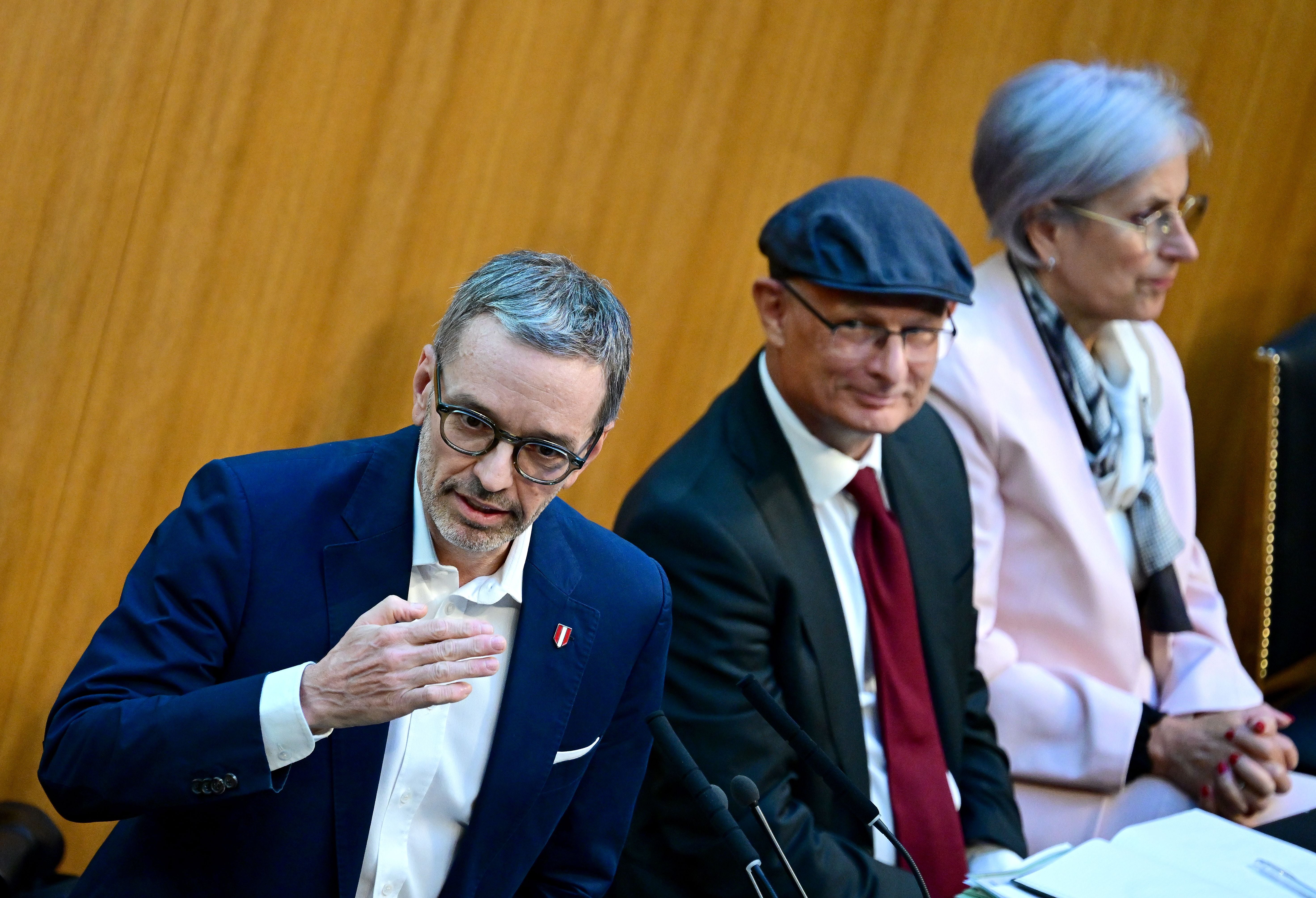 Drei Personen sitzen im Parlament, eine Person spricht an einem Mikrofon.