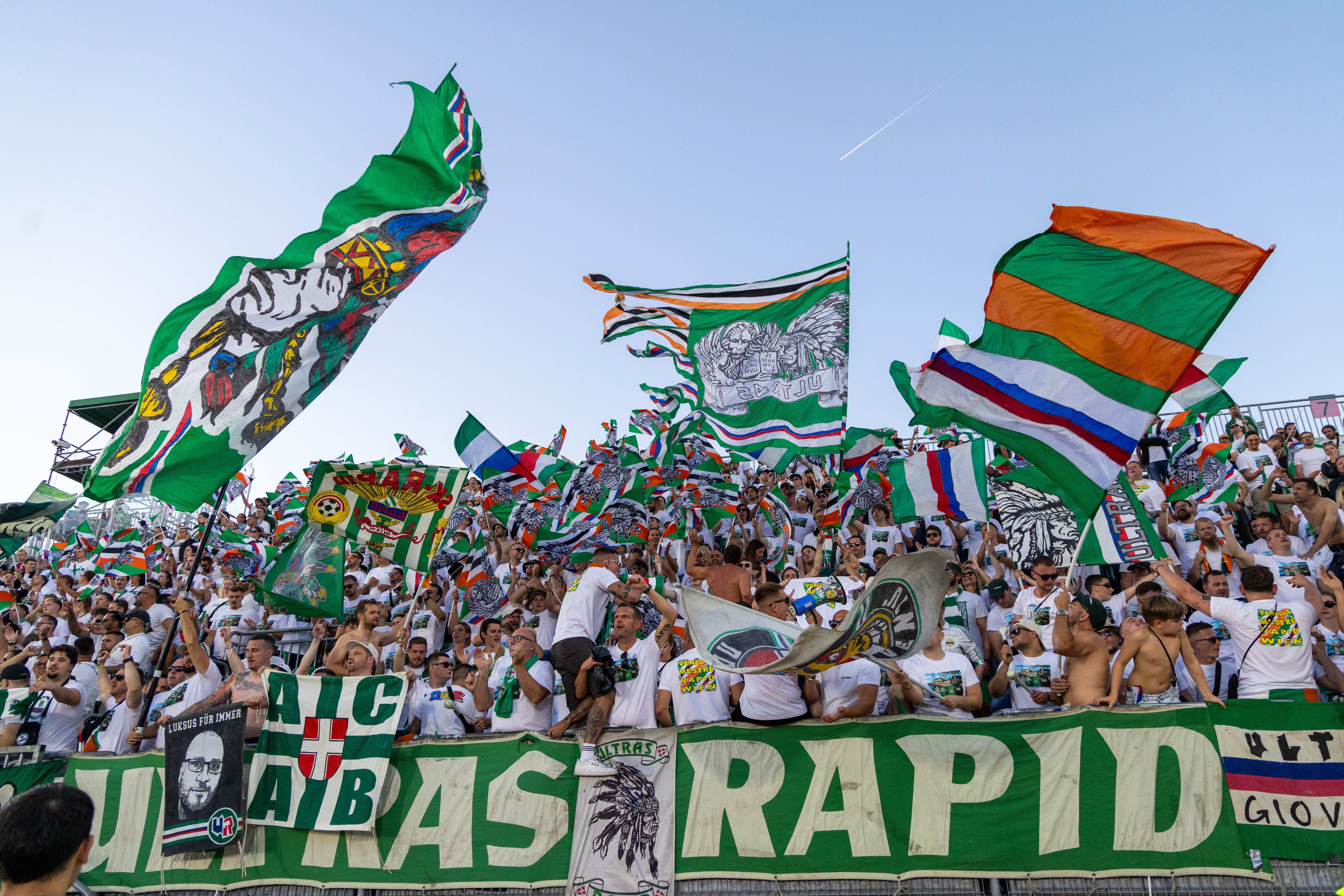 Rapid-Fans jubeln im Stadion, schwenken grüne Fahnen und tragen weiße T-Shirts.