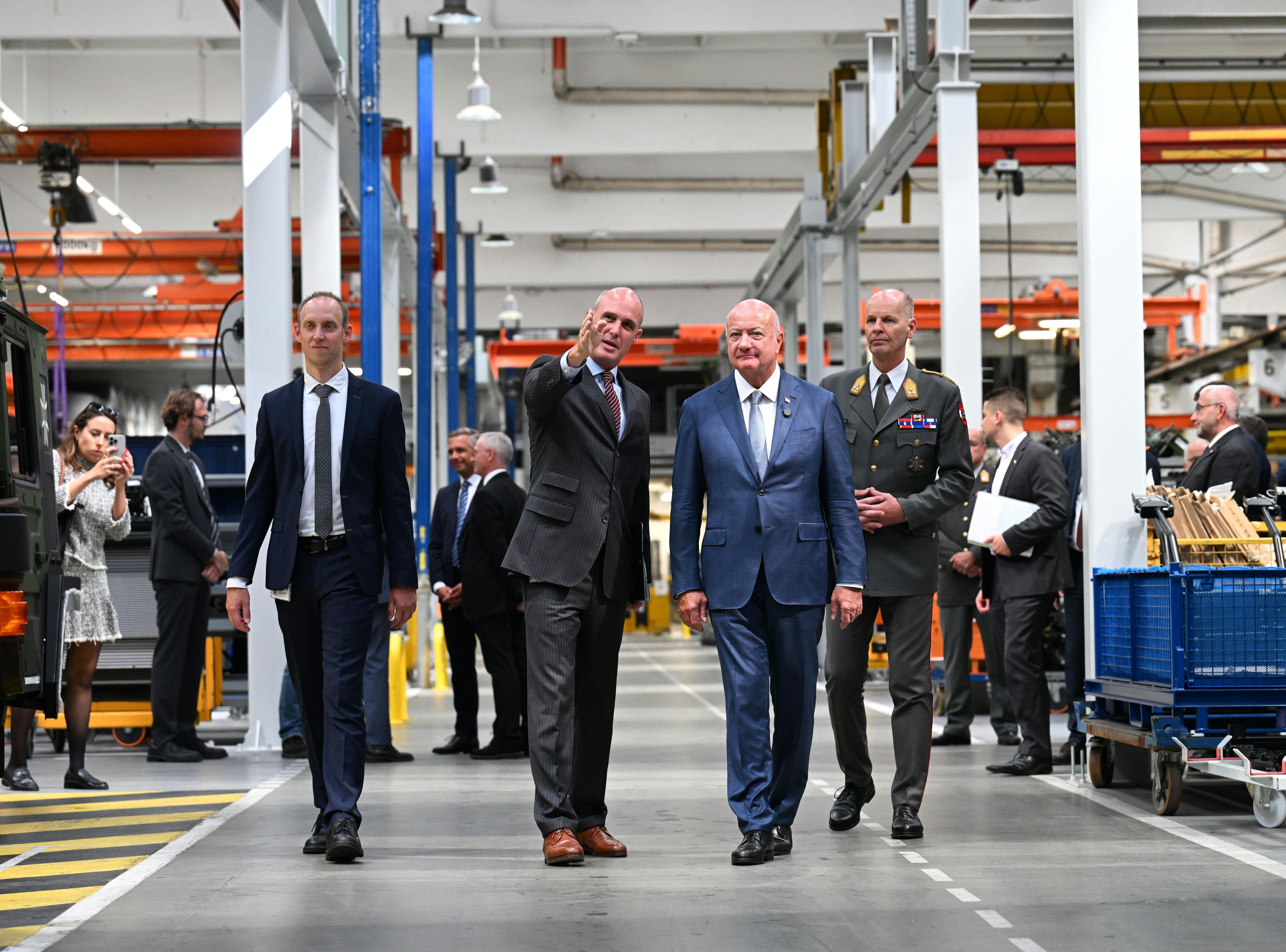 Mehrere Männer in Anzügen und Uniformen gehen durch eine moderne Fabrikhalle.