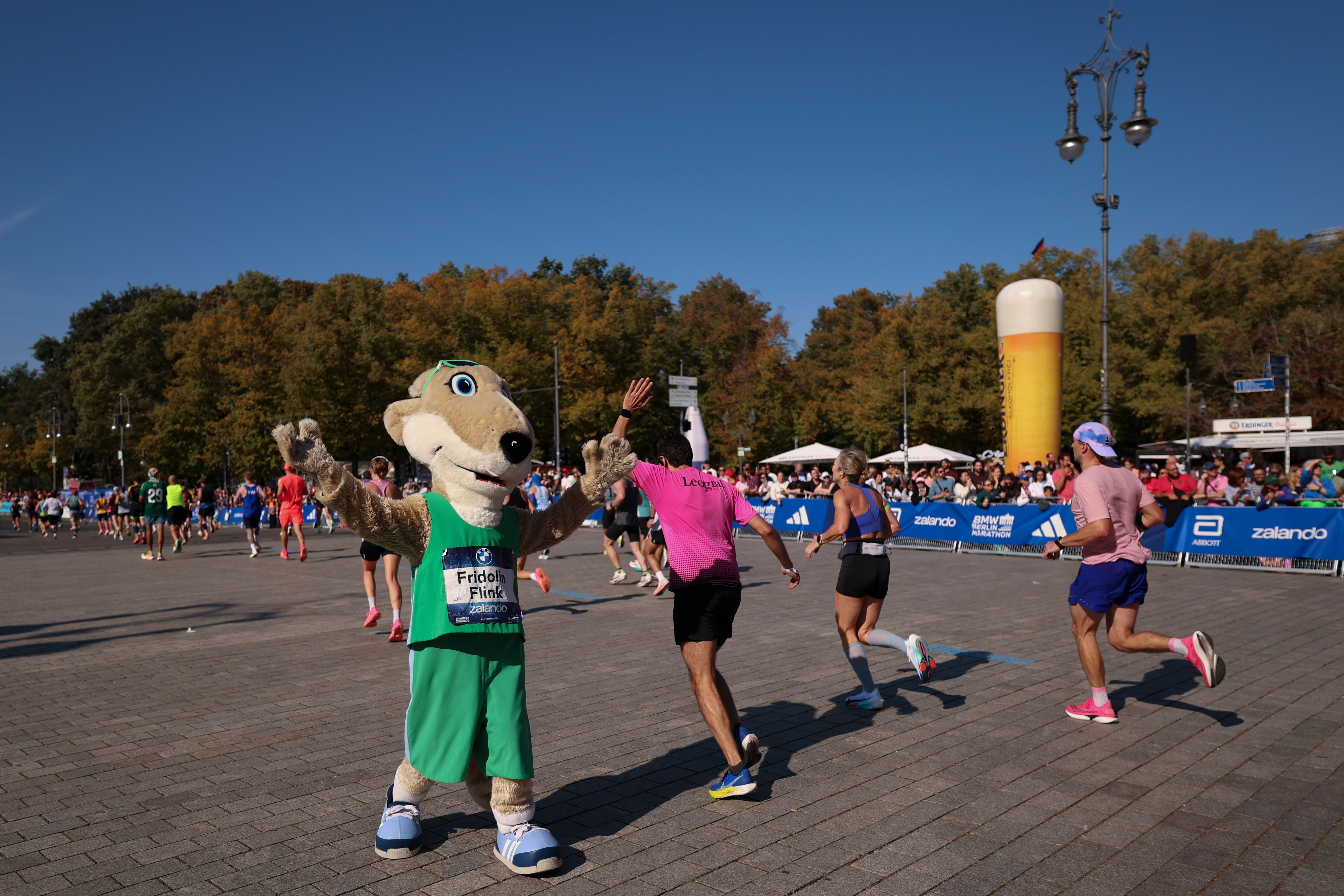 Ein Maskottchen begrüßt Läufer beim Berlin-Marathon, Zuschauer und herbstliche Bäume im Hintergrund.