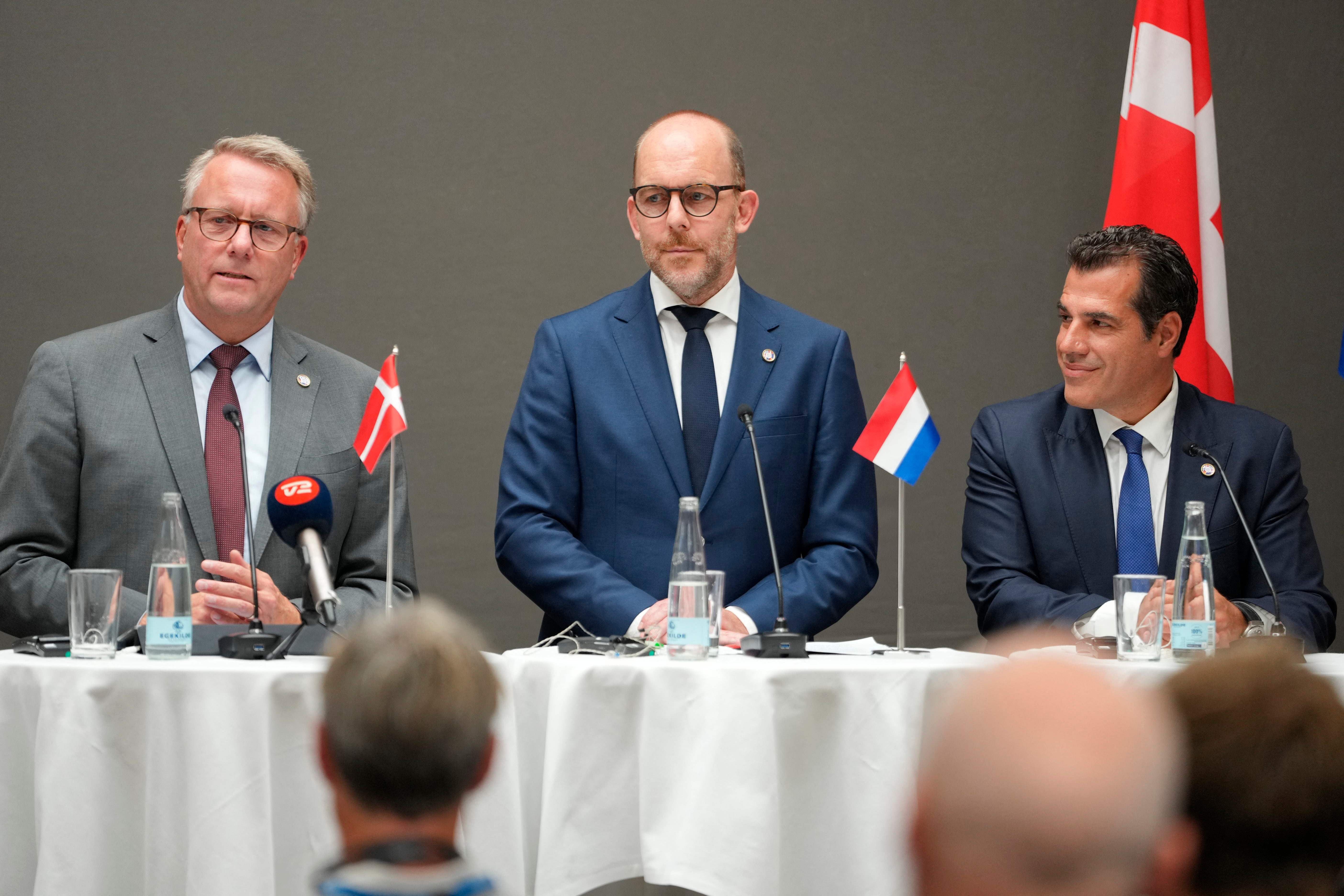 Drei Männer stehen bei einer Pressekonferenz mit Landesflaggen und Mikrofonen auf einem Tisch.