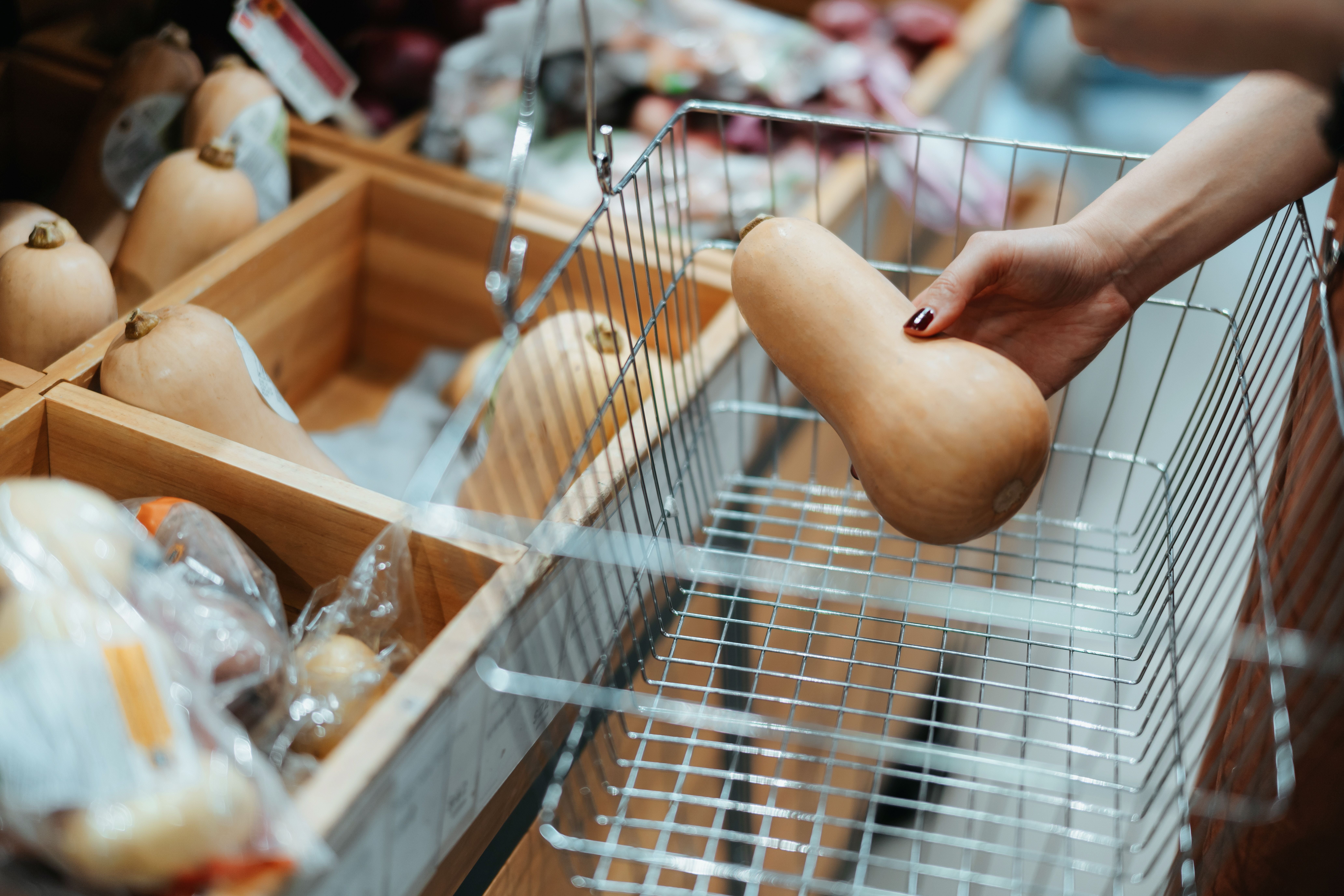 Eine Hand legt einen Butternut-Kürbis in einen leeren Einkaufskorb im Supermarkt.