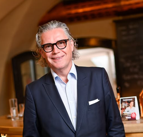 Ein Mann mit Brille und Anzug steht lächelnd in einem eleganten Innenraum.
