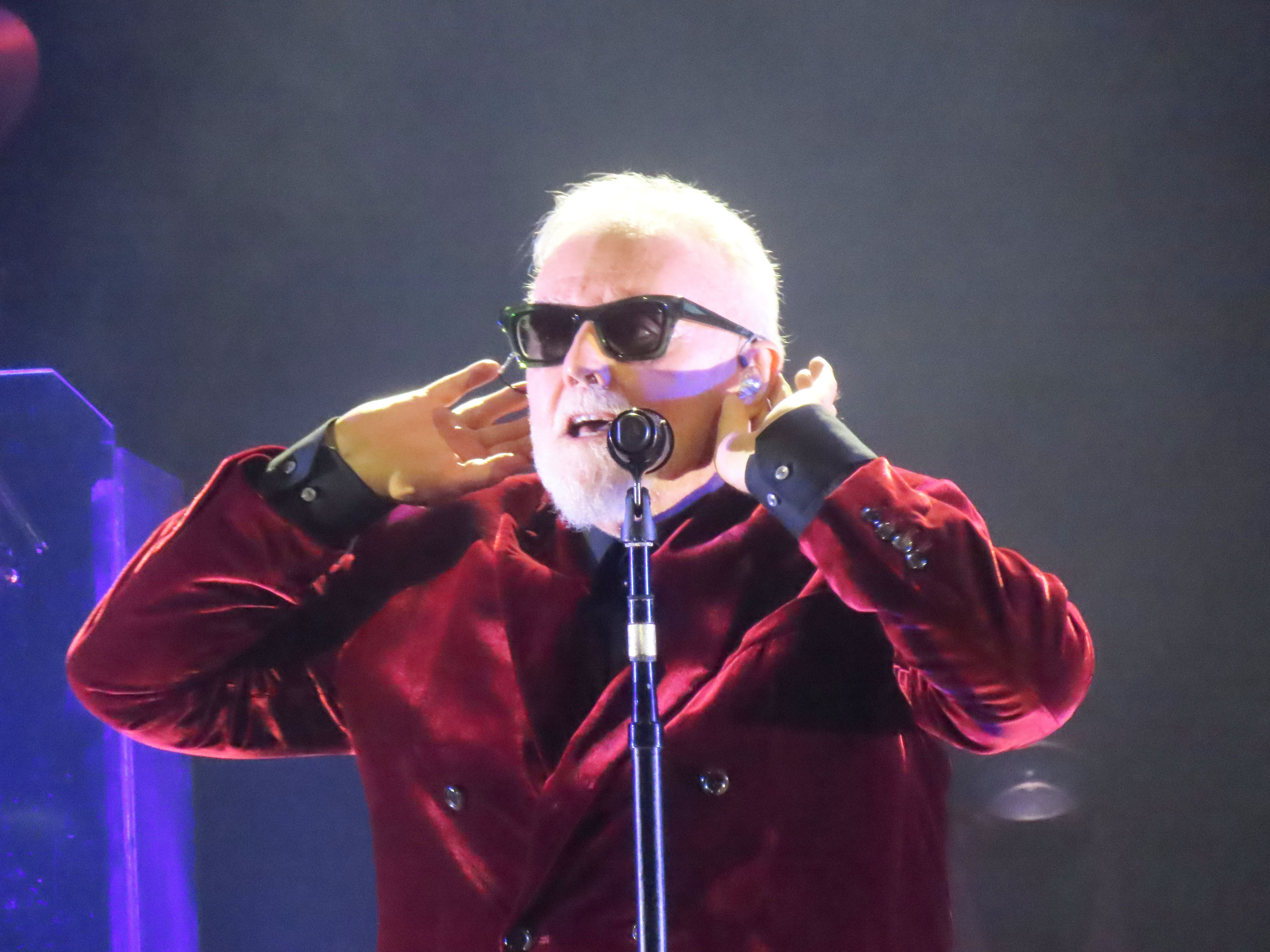 Älterer Mann mit Sonnenbrille und rotem Samtblazer singt auf einer Bühne ins Mikrofon.