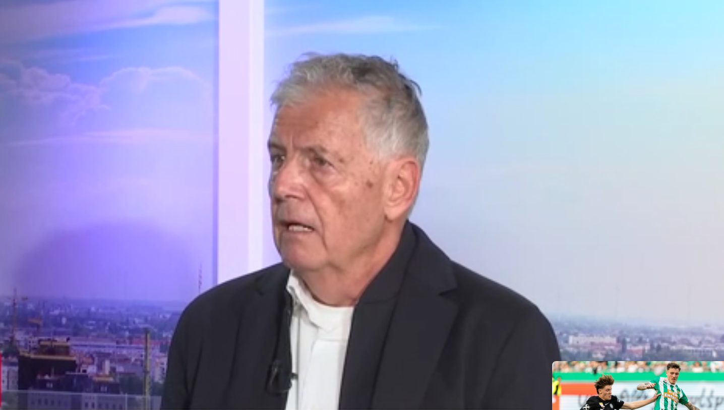 Ein älterer Mann im Anzug spricht in einem Fernsehstudio mit Stadtpanorama im Hintergrund.