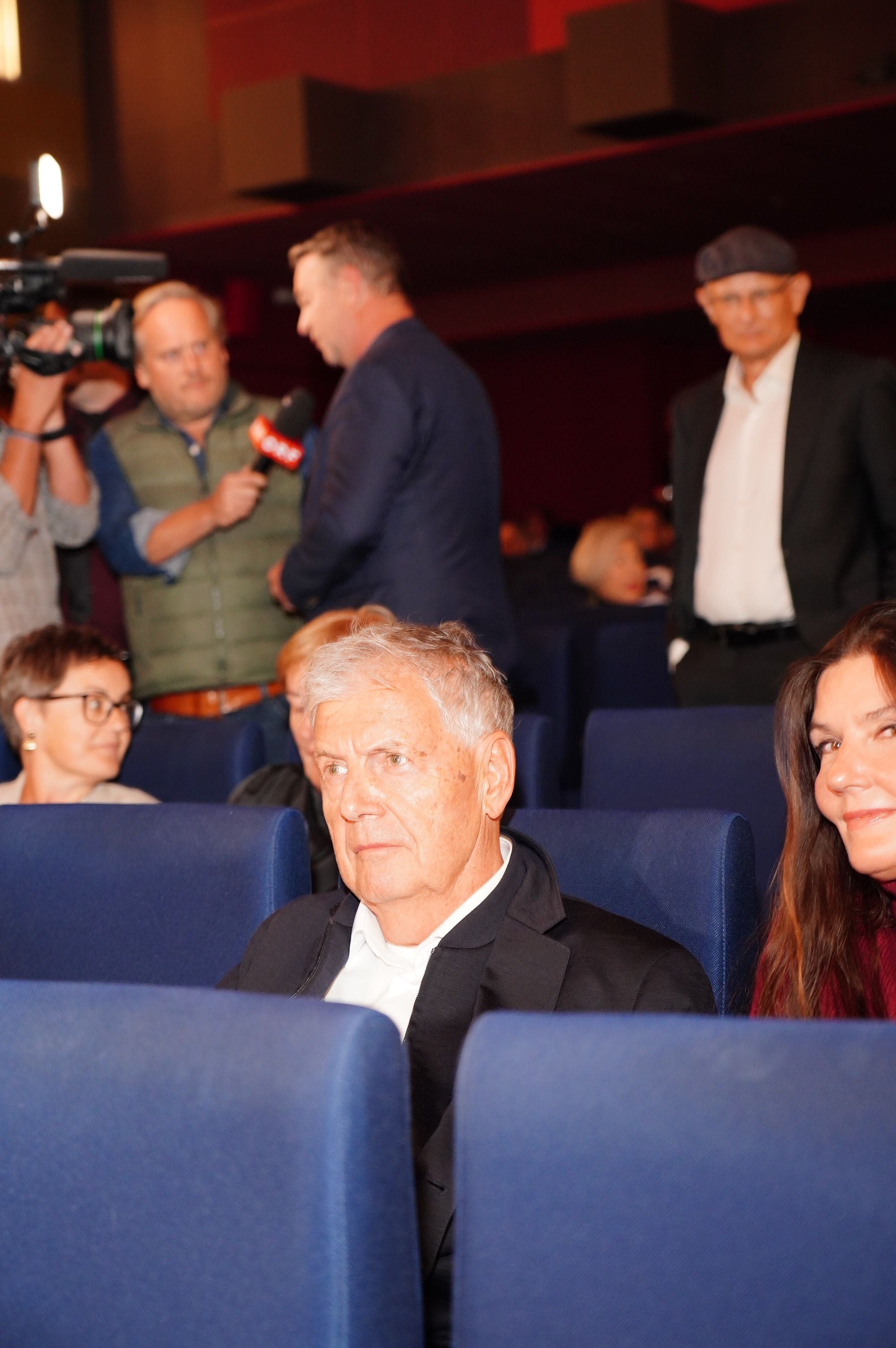 Mehrere Personen sitzen und stehen in einem Kinosaal, im Vordergrund ein älterer Mann in schwarzen Jackett.