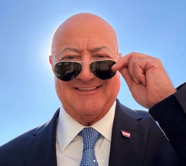Ein älterer Mann im Anzug hebt seine Sonnenbrille vor blauem Himmel leicht an.