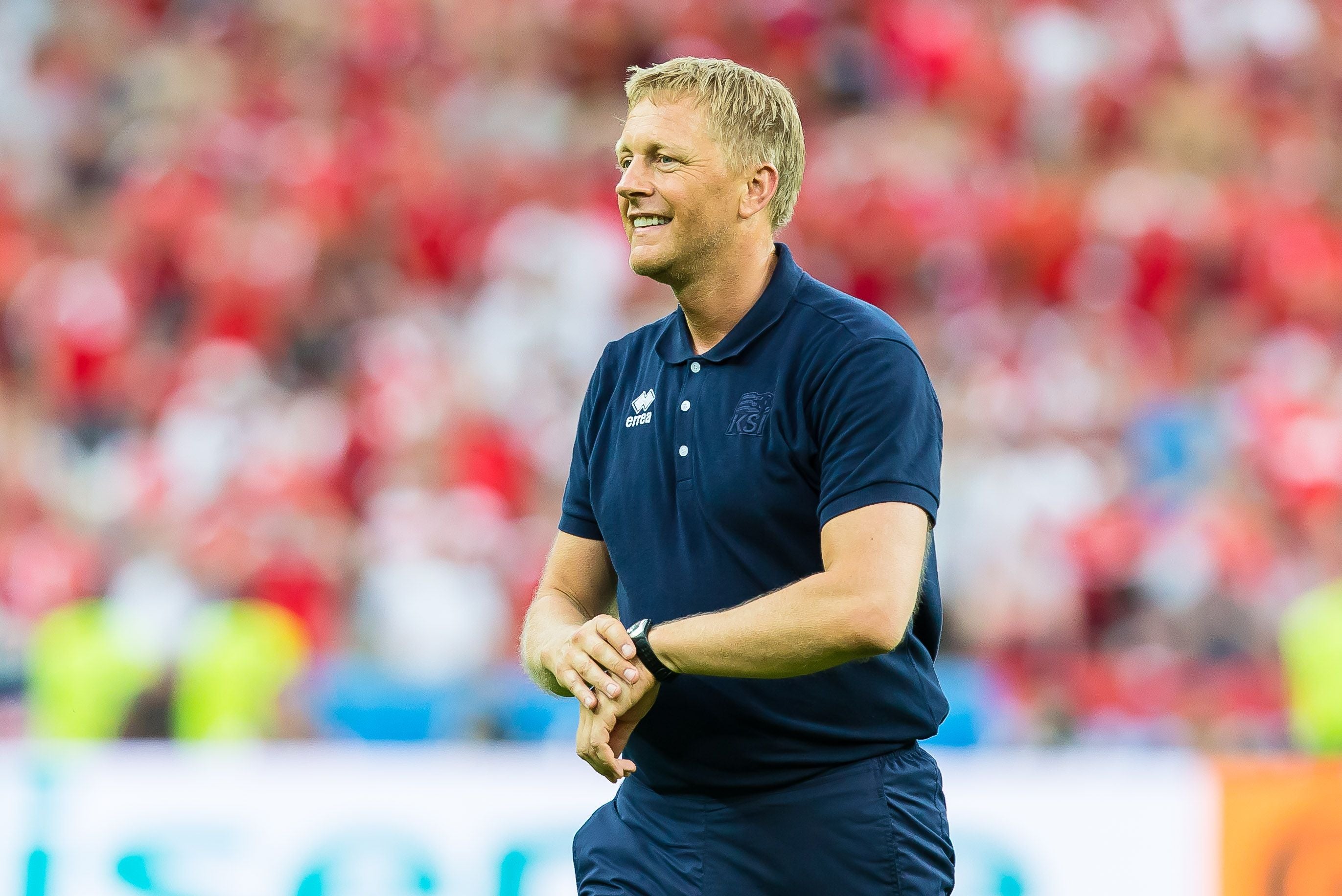 Ein lächelnder Fußballtrainer im blauen Poloshirt auf dem Spielfeld bei einem Spiel.