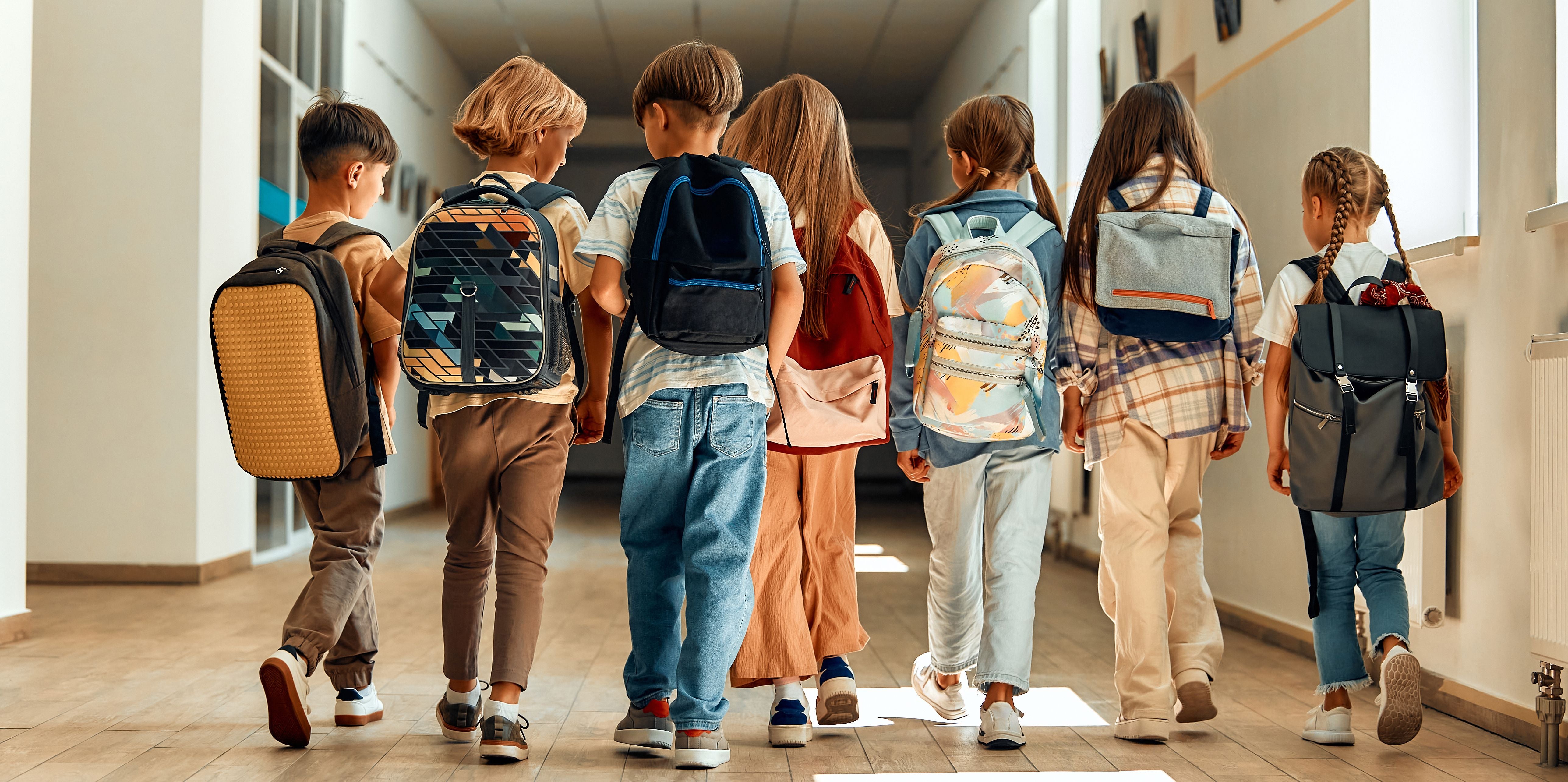 Sieben Schulkinder mit Rucksäcken gehen gemeinsam durch einen hellen Schulflur.