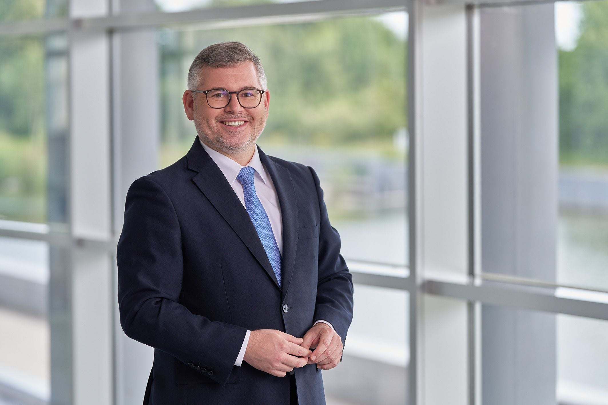 Ein lächelnder Mann im Anzug steht vor großen Fenstern mit Blick ins Grüne.