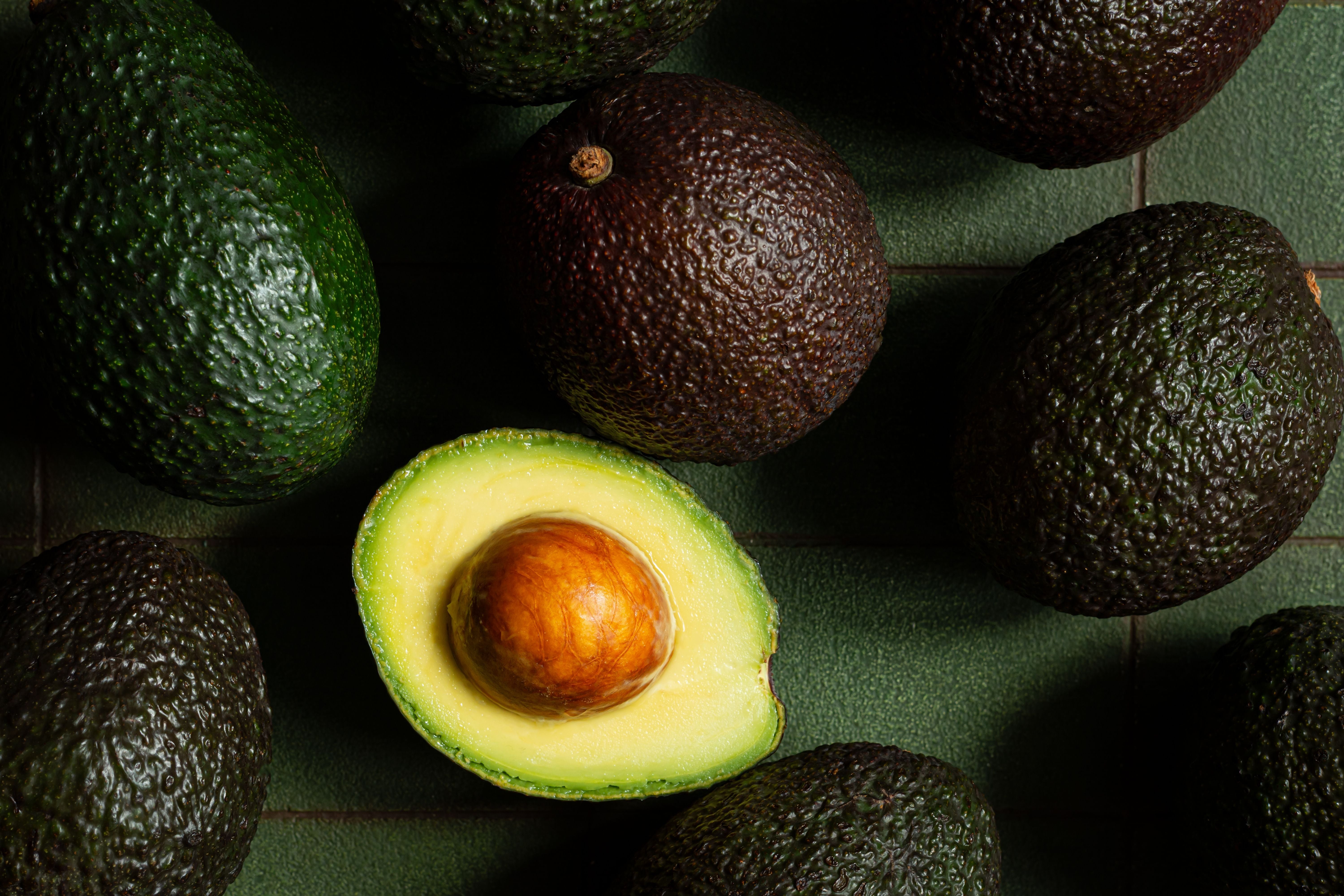 Mehrere reife Avocados, eine davon halbiert, auf dunkelgrünem Untergrund.