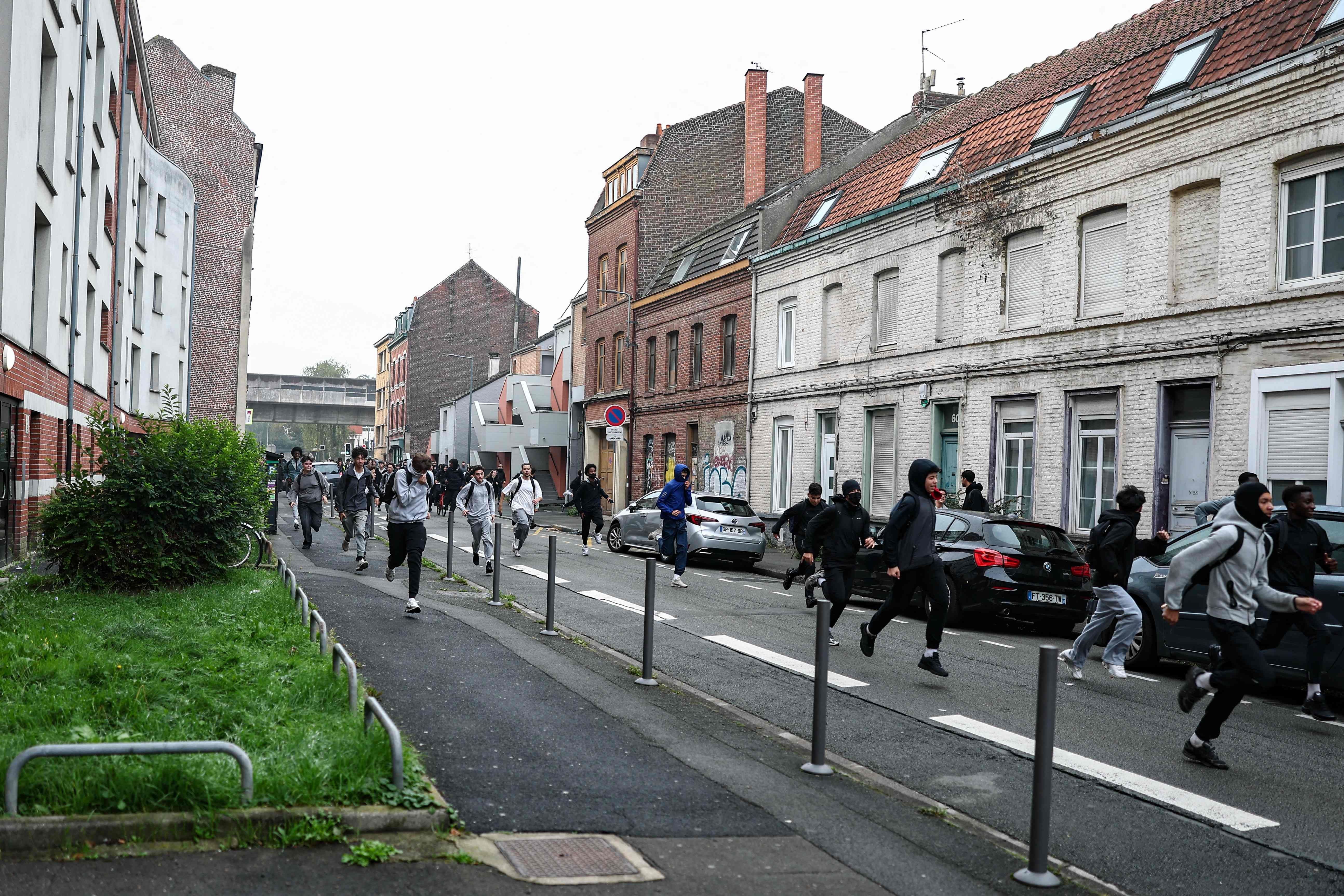 Mehrere Menschen rennen auf einer Straße in einer Stadt, einige tragen Kapuzen und Rucksäcke.