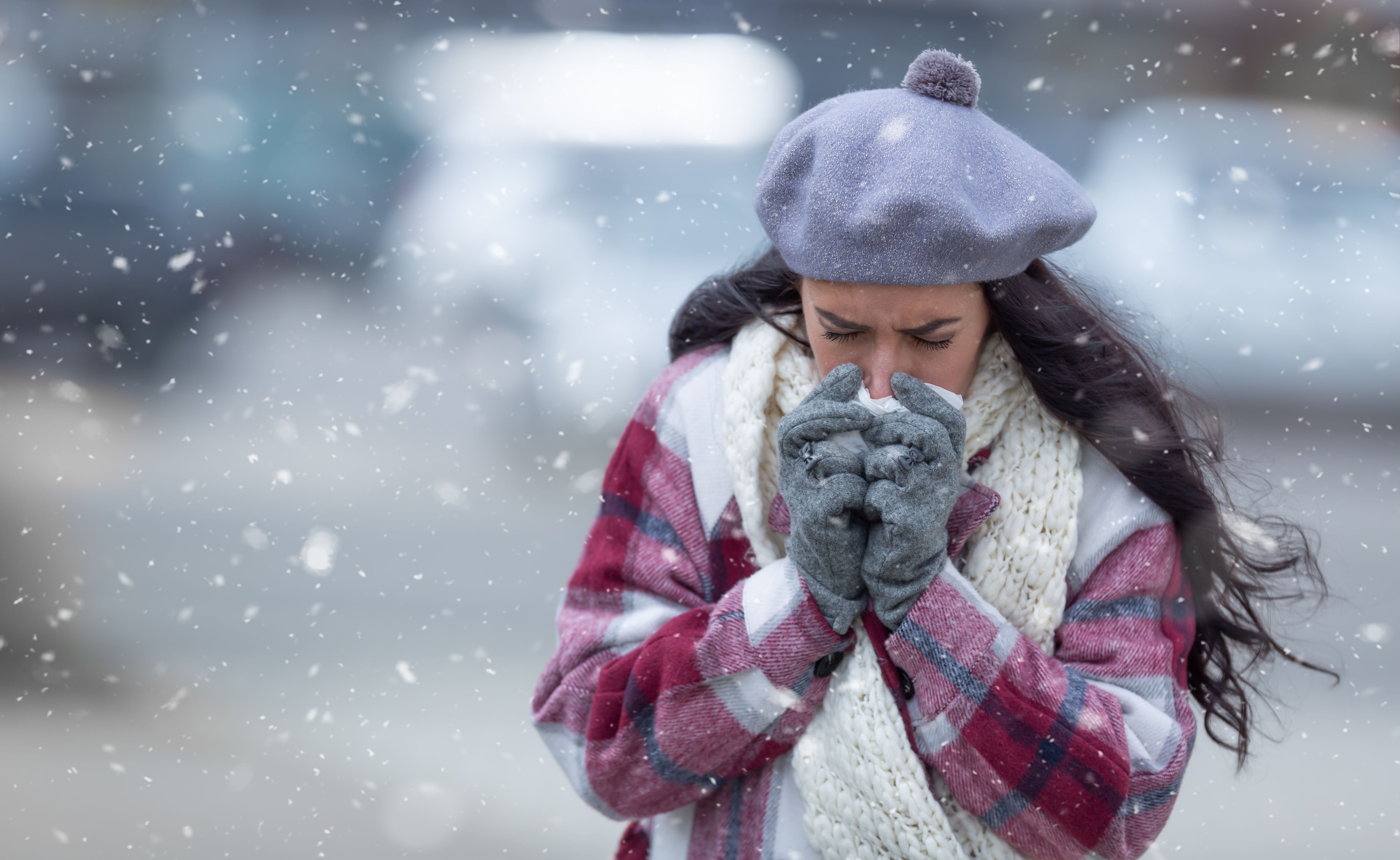 Frau putzt sich bei Schneefall draußen die Nase und trägt Winterkleidung.