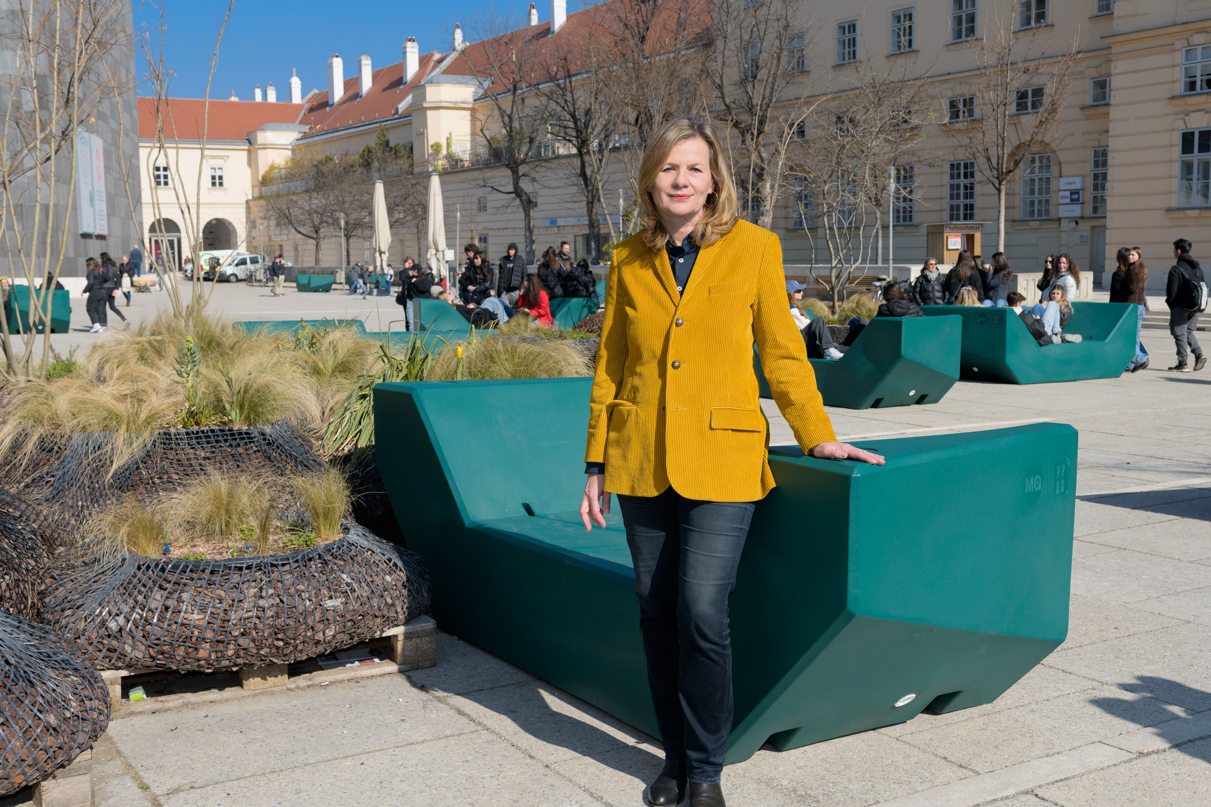 Frau im gelben Blazer steht an einer grünen Bank im sonnigen Museumsquartier.