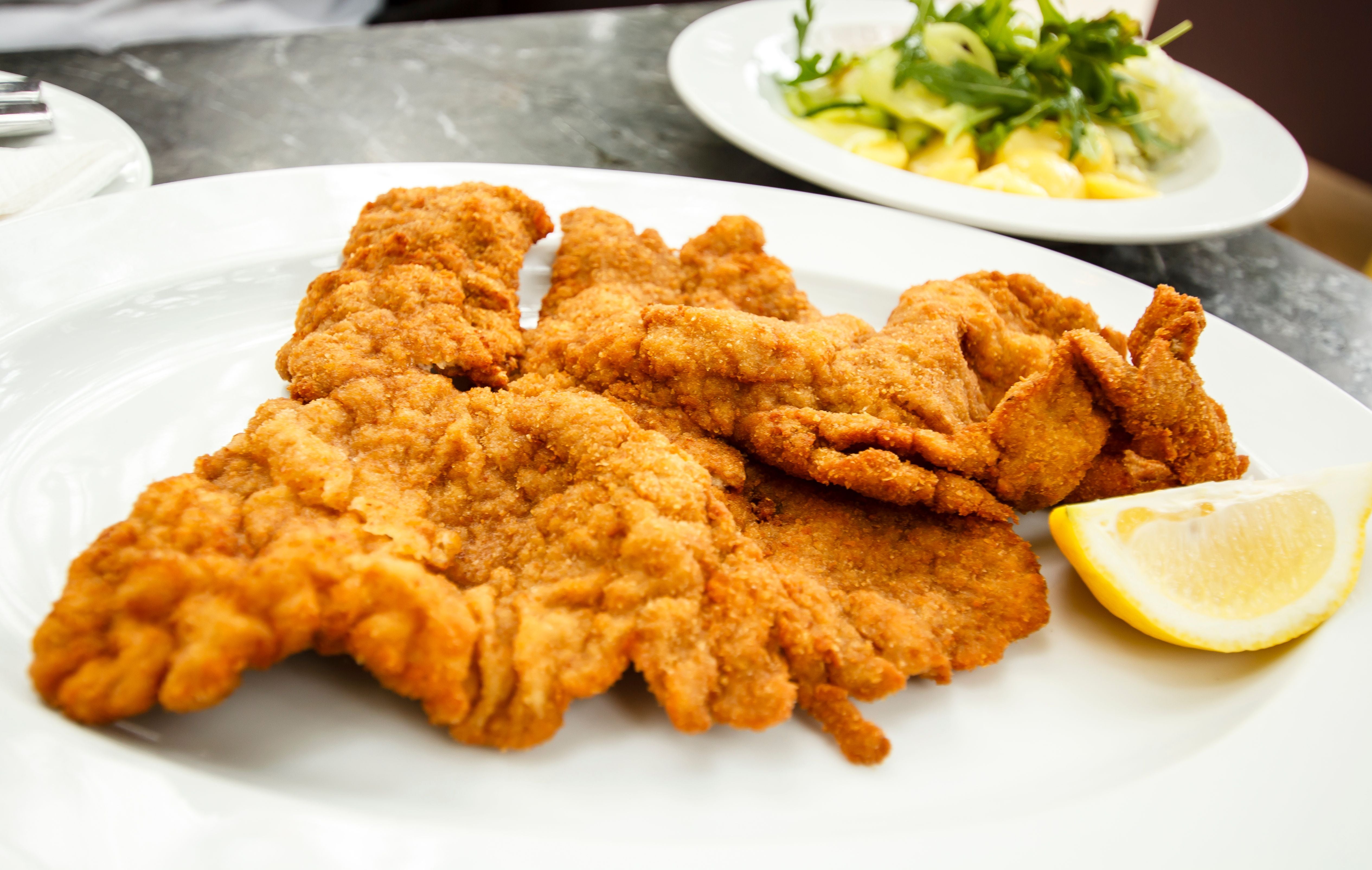 Wiener Schnitzel mit Zitronenscheibe auf weißem Teller, im Hintergrund ein Salatteller.