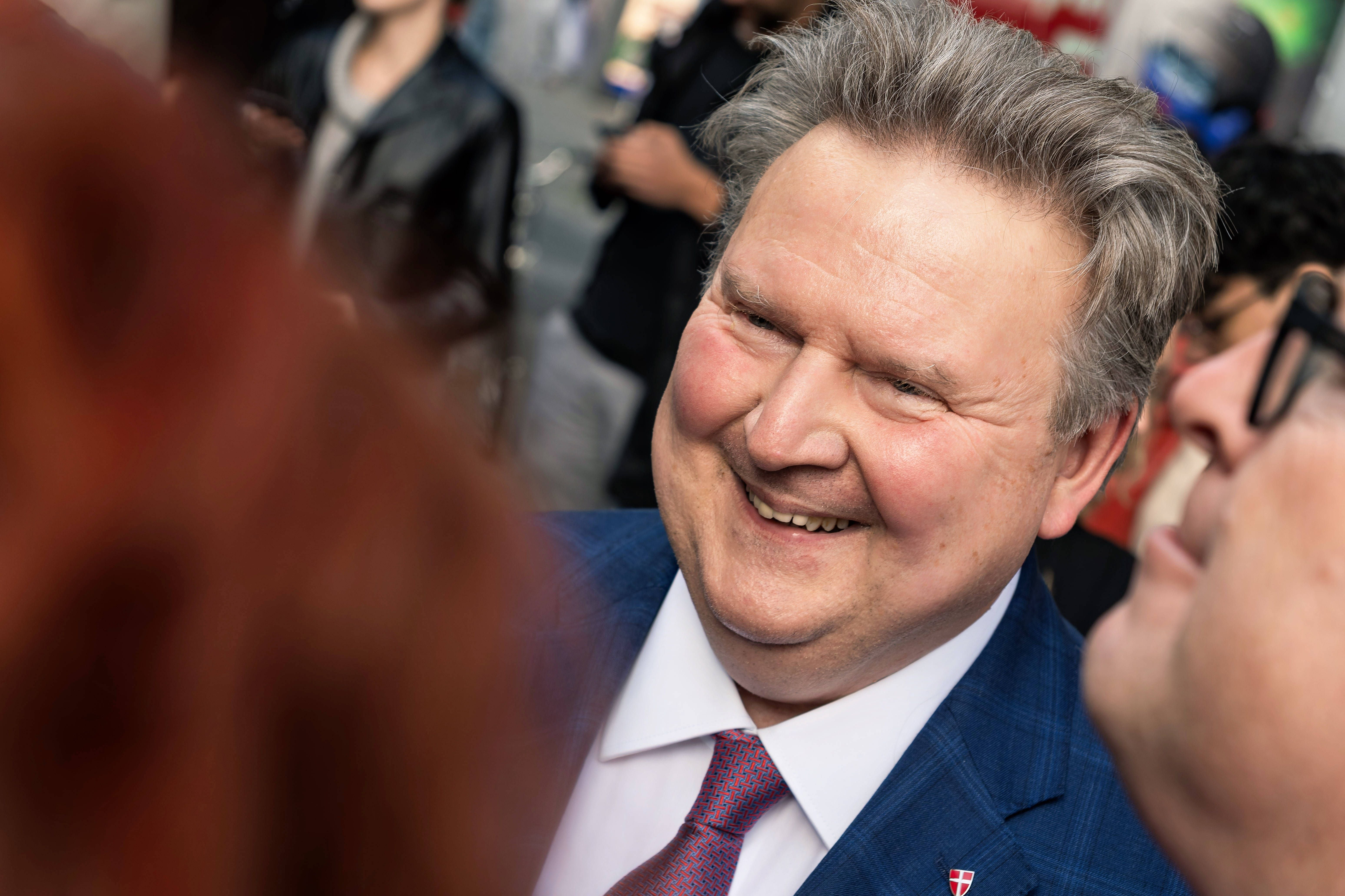 Ein lächelnder Mann im Anzug auf einer Wahlkampfveranstaltung im Freien, umgeben von Menschen.