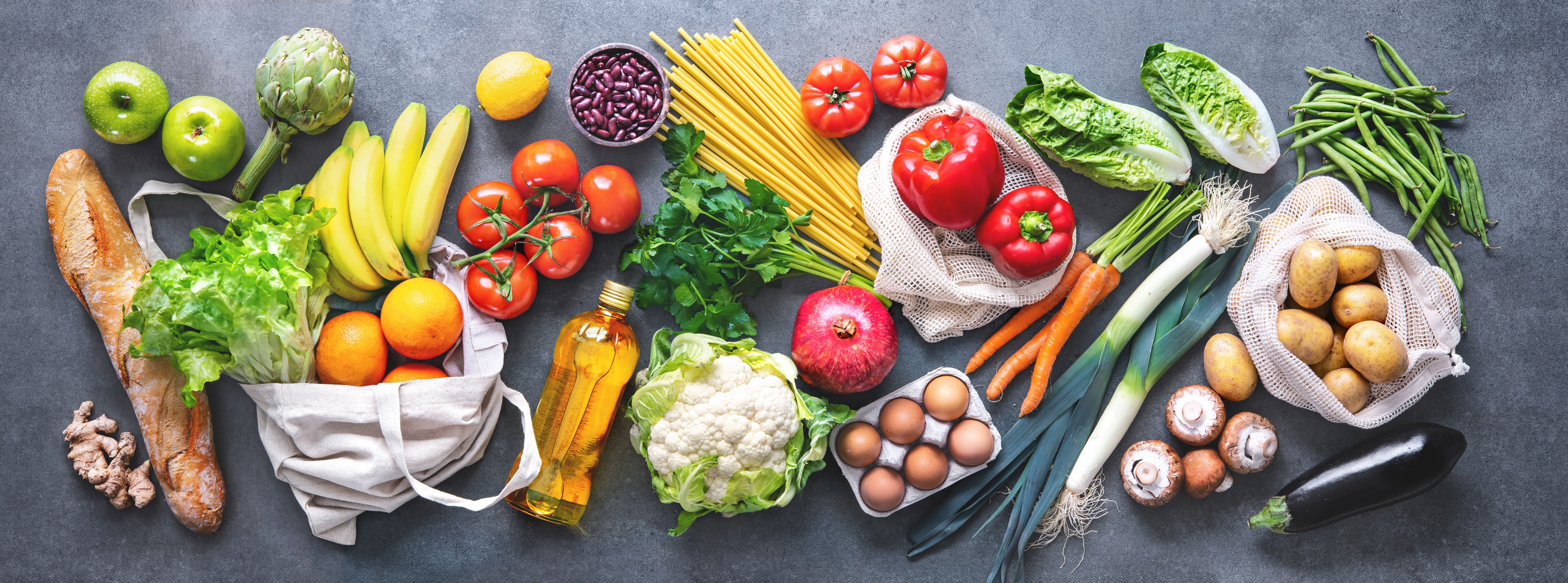 Obst, Gemüse, Brot und Öl in wiederverwendbaren Beuteln auf einer grauen Oberfläche.